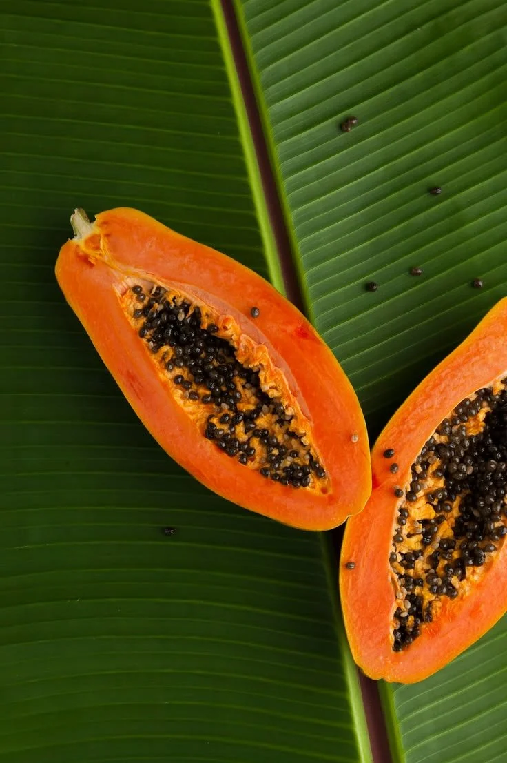 Close-up of two halves of a ripe papaya fruit with orange flesh and black seeds on large green tropical leaves.
