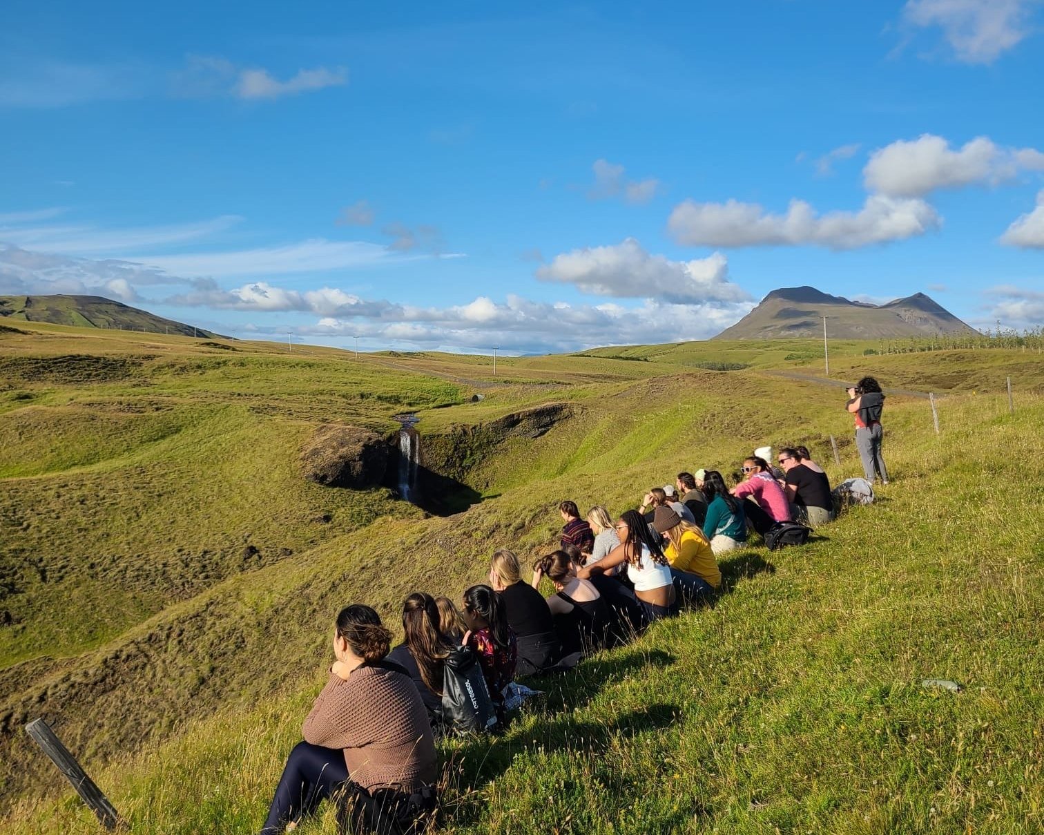 Group of people sitting on grassy hillside overlooking a landscape with a small waterfall, mountains in the distance, and a blue sky with scattered clouds.