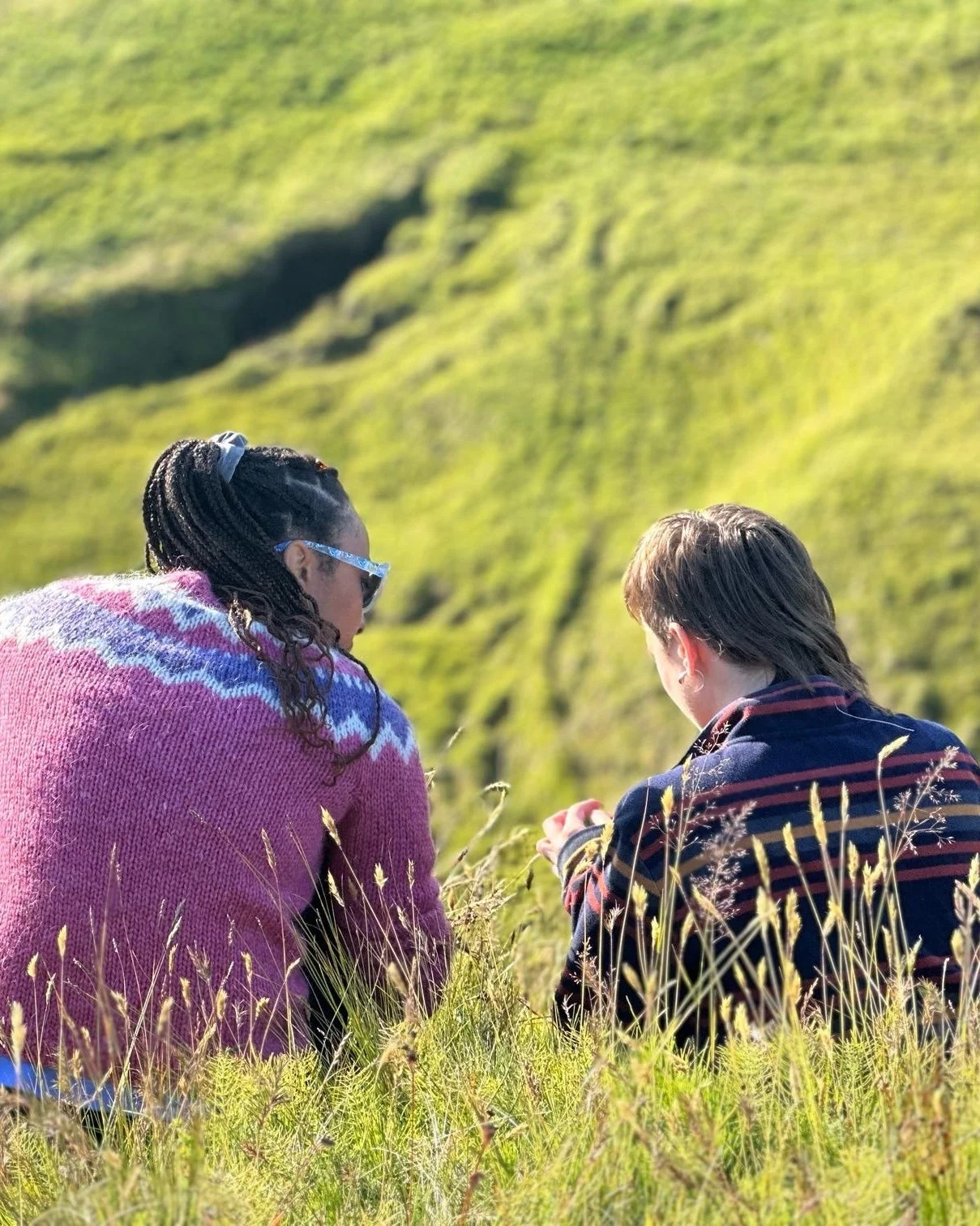Two women sitting outdoors in a grassy field with a green hill in the background, one wearing a pink sweater with a colorful pattern and sunglasses, the other in a striped jacket, both looking at something in their hands.