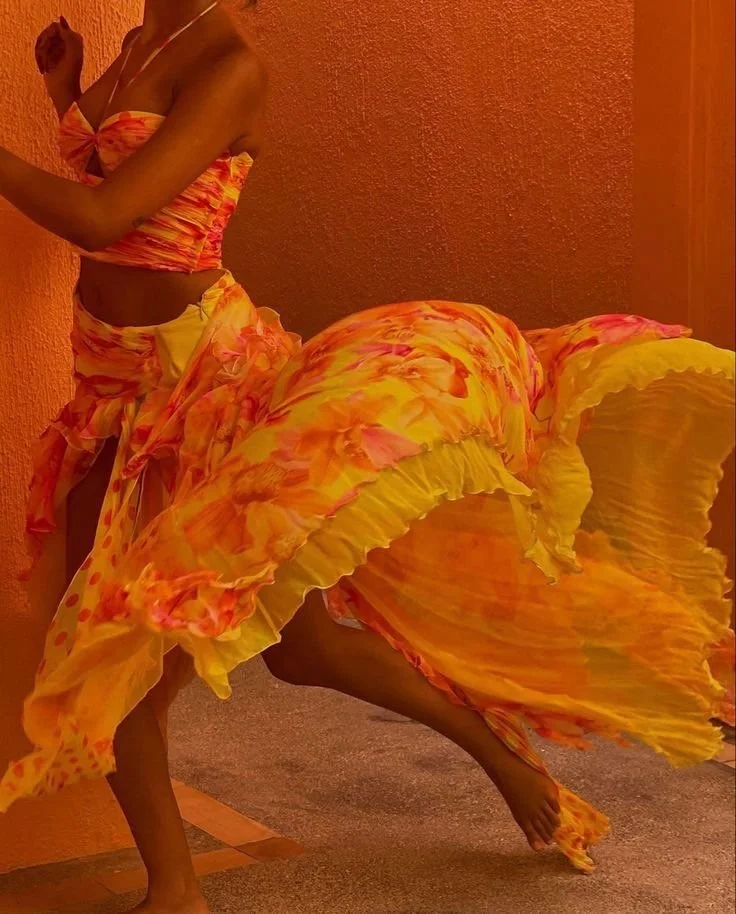 Person dancing in a flowing orange and yellow dress with pink accents, standing against a textured orange wall.