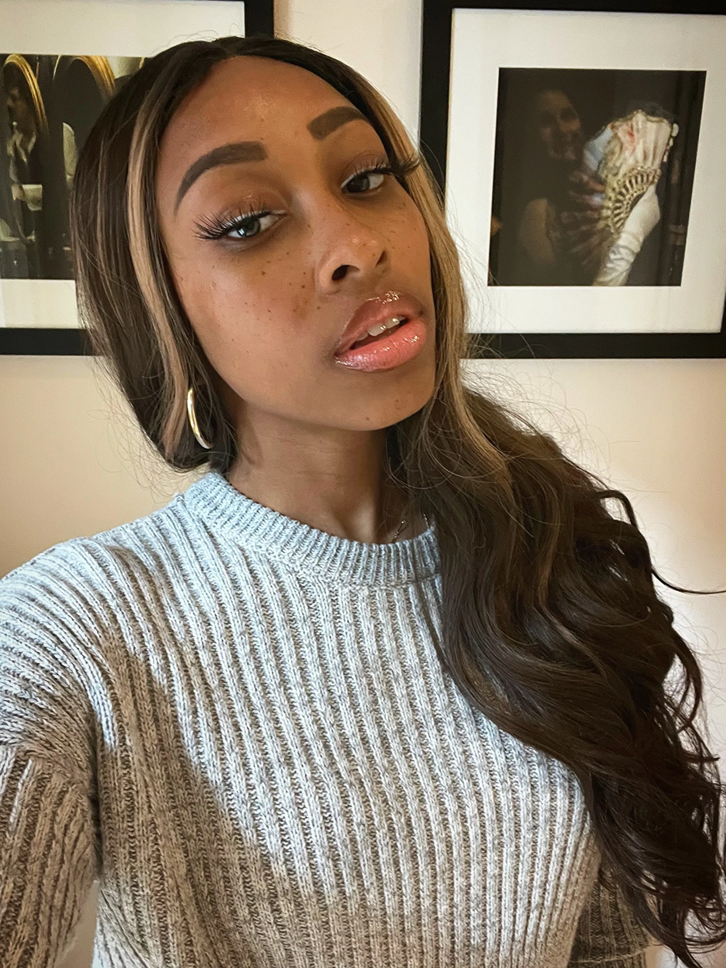 A young woman with long, wavy brown hair and brown skin. She's wearing a light gray, ribbed, knit sweater and gold hoop earrings, standing in front of framed artwork on a beige wall.