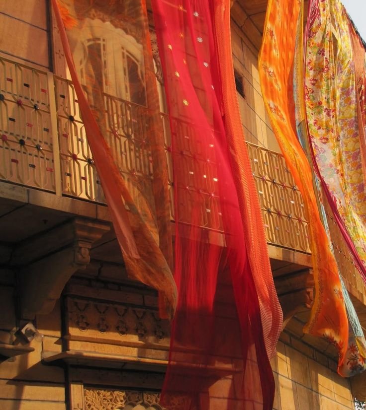 Colorful fabrics or curtains hanging outside on a balcony, with a building facade and ornate architectural details in the background.