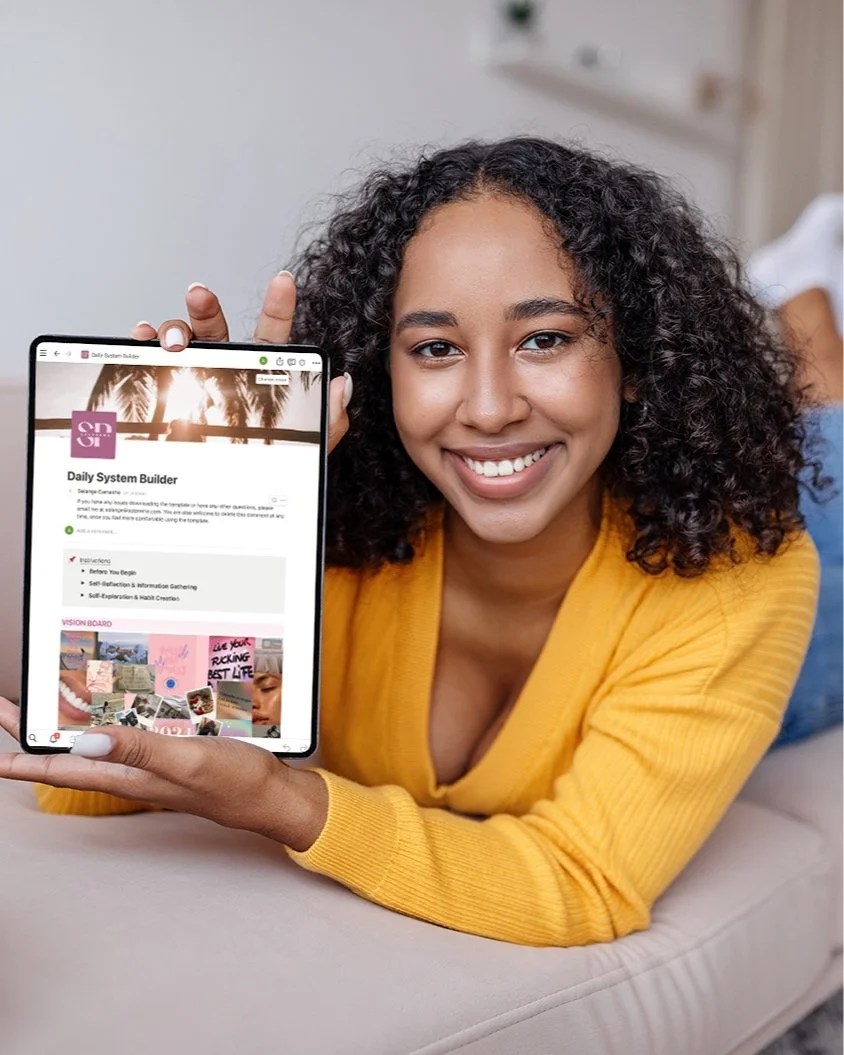 A woman with curly hair smiling and lying on her stomach on a light-colored couch, holding a tablet displaying a webpage titled 'Daily System Builder'.