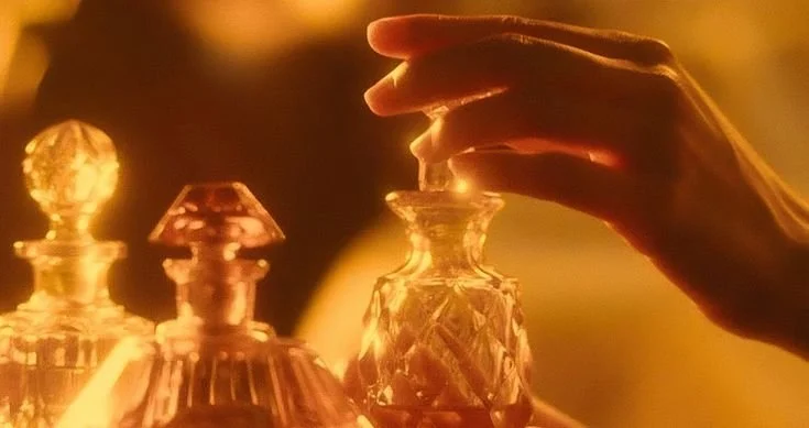 A hand reaching towards a collection of ornate perfume bottles with a warm, golden glow.