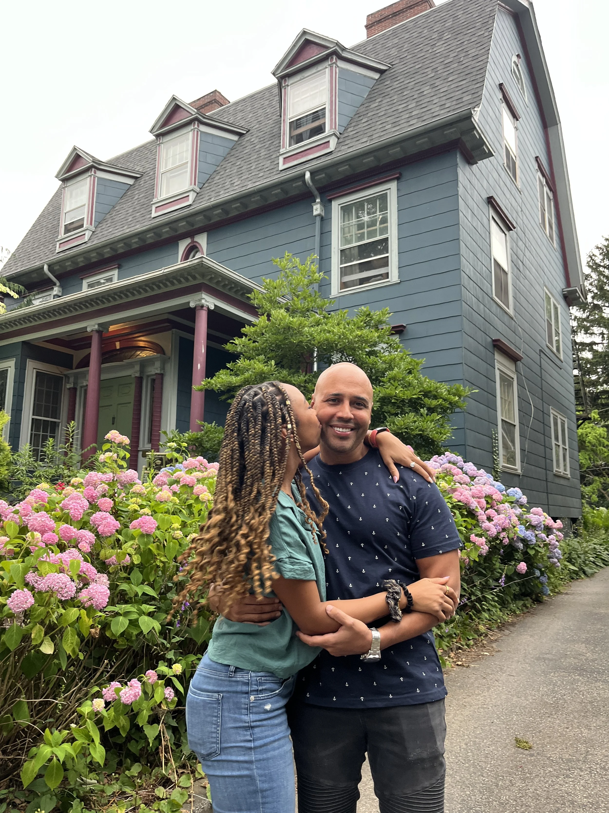 A woman and man embracing and smiling in front of a blue house with pink and white trim, surrounded by pink and purple hydrangea flowers.