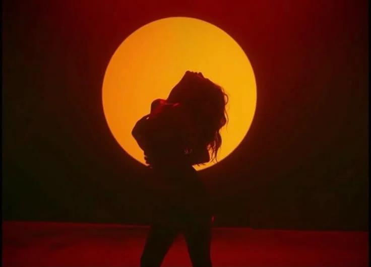Person posing in front of a large yellow circle resembling the sun, with their hand on their head and long hair, in a dark setting with a red floor.