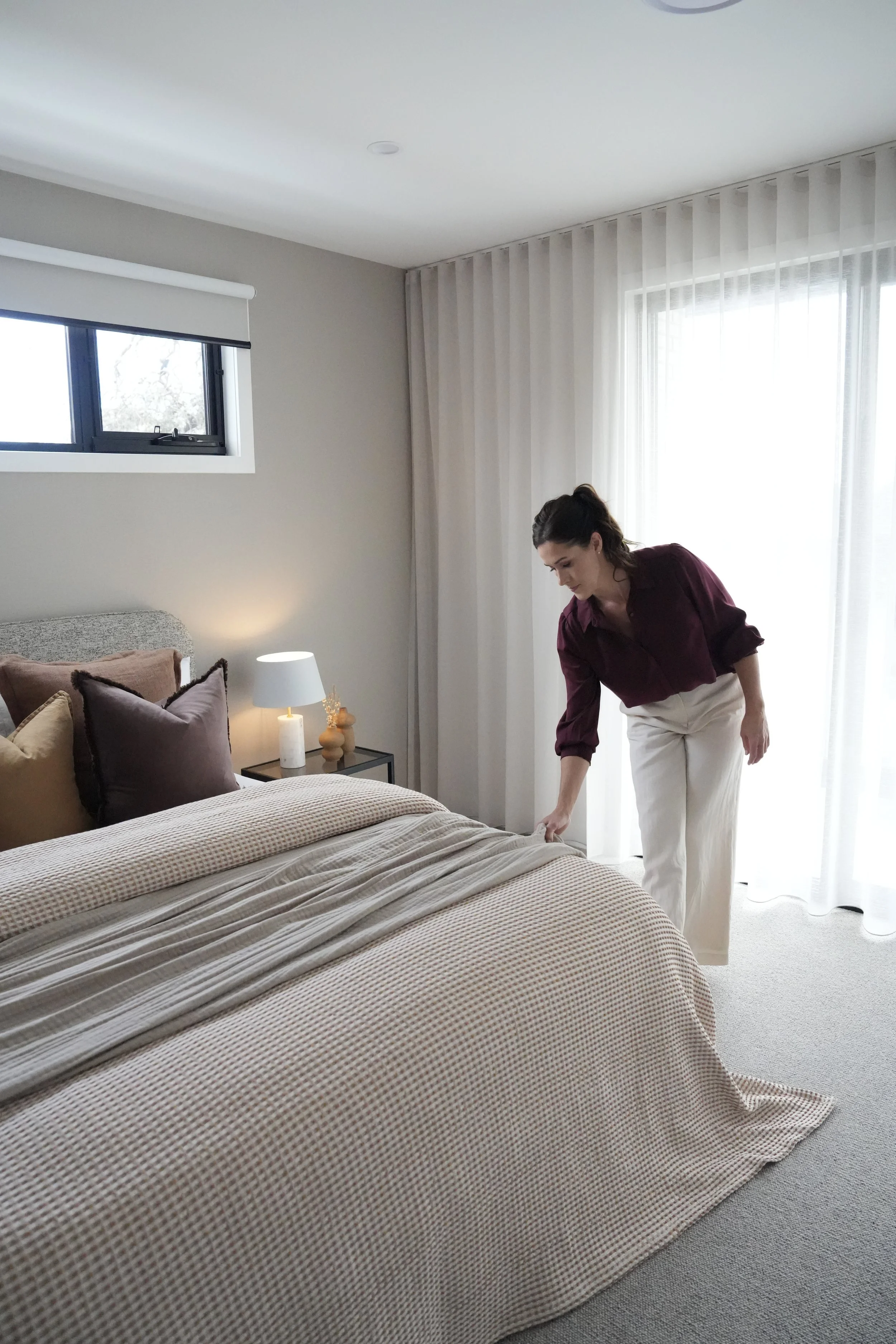 Woman making the bed in a bedroom with beige and brown pillows, a white lamp on a black bedside table, and sheer white curtains.