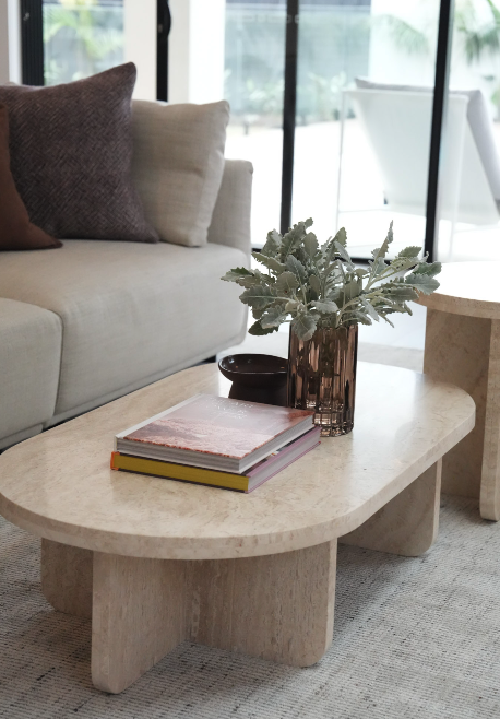 Living room with a light-colored sofa, purple and beige pillows, a marble coffee table with books and a plant in a vase, and a sliding glass door leading outside.