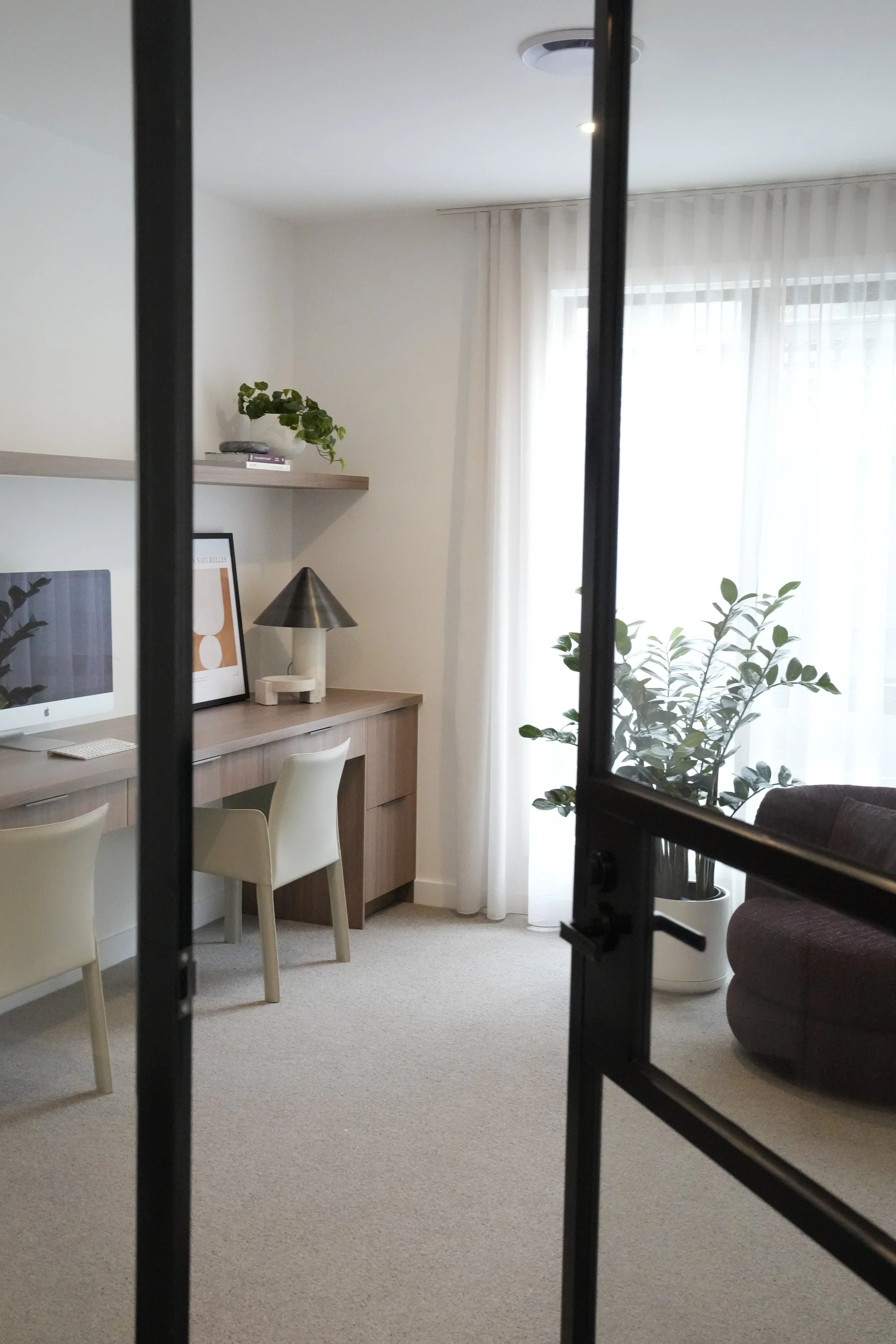 Living room with a desk, chairs, a computer, framed artwork, a lamp, potted plants, and sheer curtains over windows.