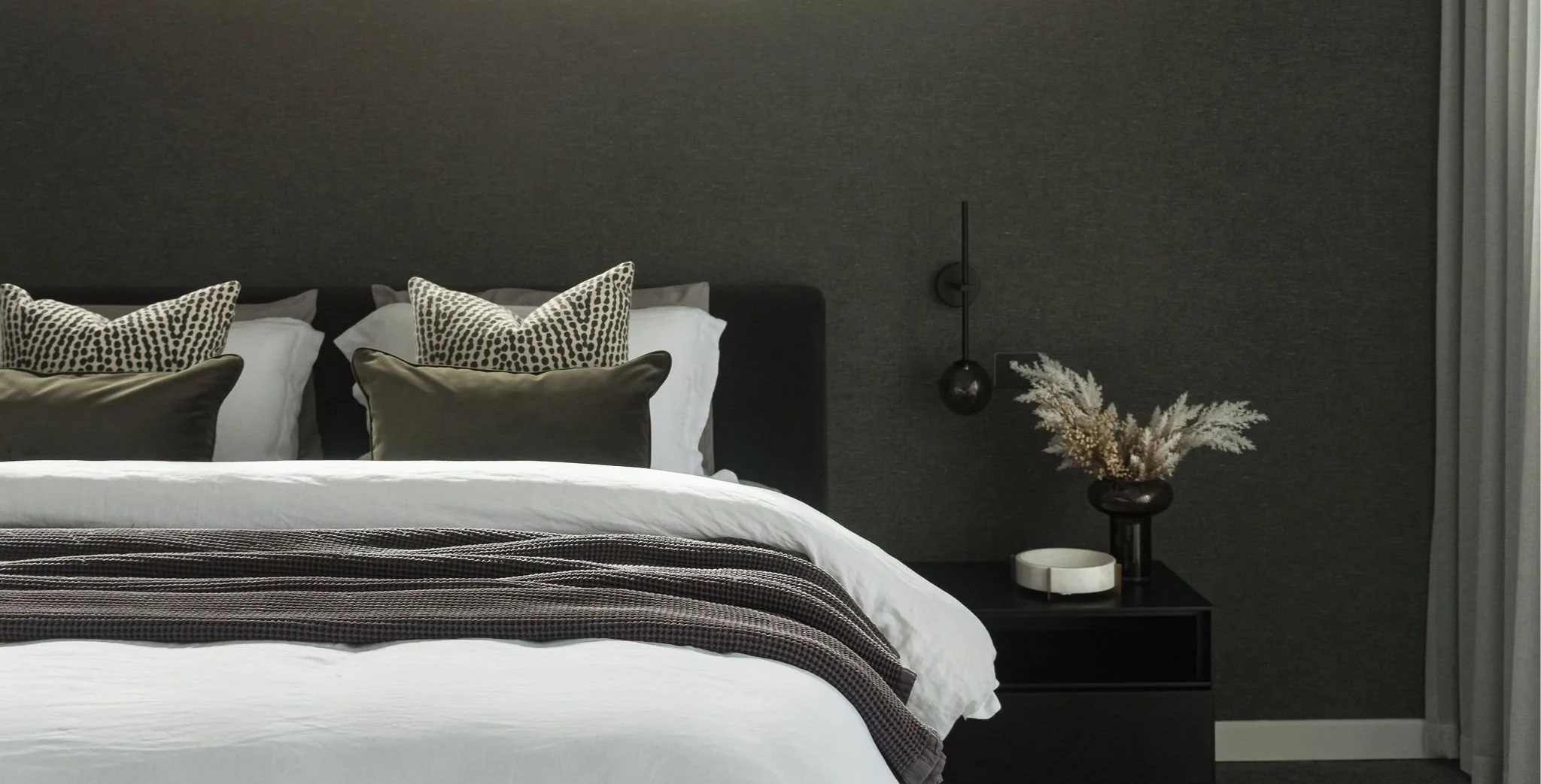 Modern bedroom with a dark fabric headboard, white bedding, and decorative pillows, next to a nightstand with a vase of dried flowers and a marble dish, with gray curtains to the right.