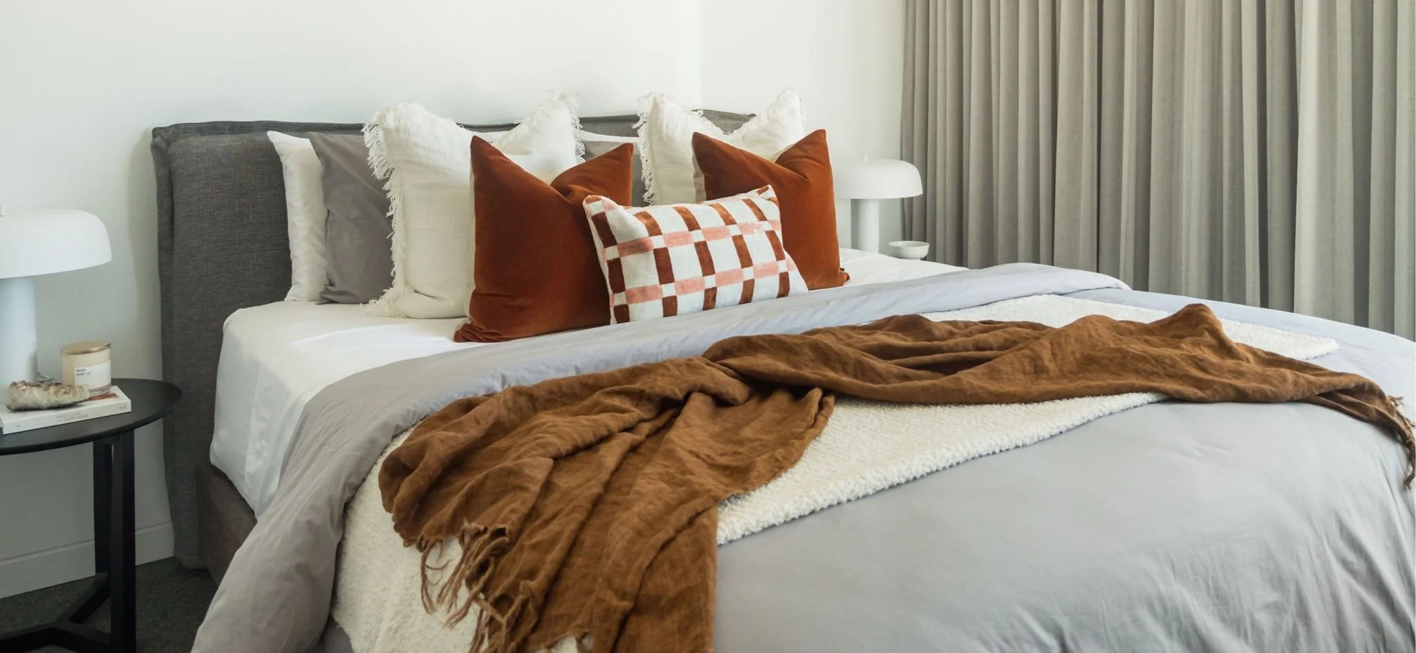 A neatly made bed with white sheet, beige blanket, and brown throw blanket. Eight pillows with various covers, including white, gray, rust, and a patterned pillow, are arranged on the bed. Two white nightstands are on either side, with lamps and smal