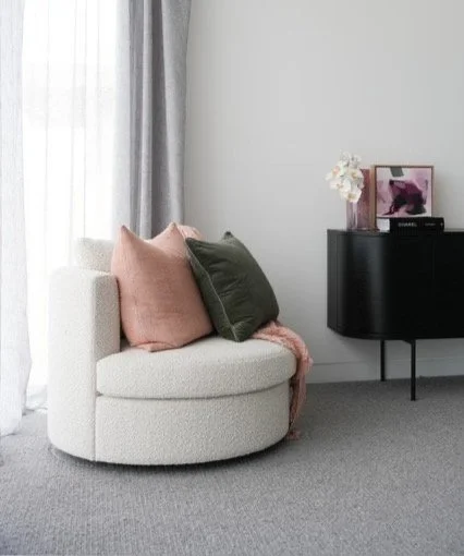 A cozy corner with a white round armchair, two pink and green cushions, a gray curtain, and a black side table with a framed picture, flowers, and a candle.
