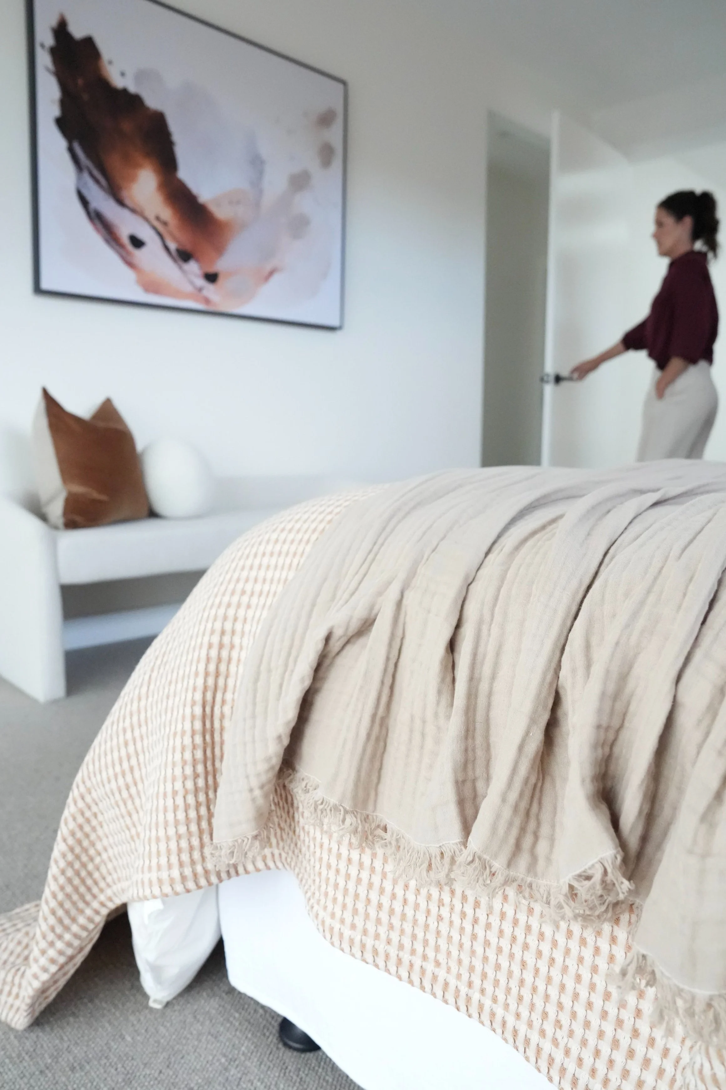 A woman opens a door in a bedroom with a bed in the foreground, a white couch with pillows, and an abstract art piece on the wall.