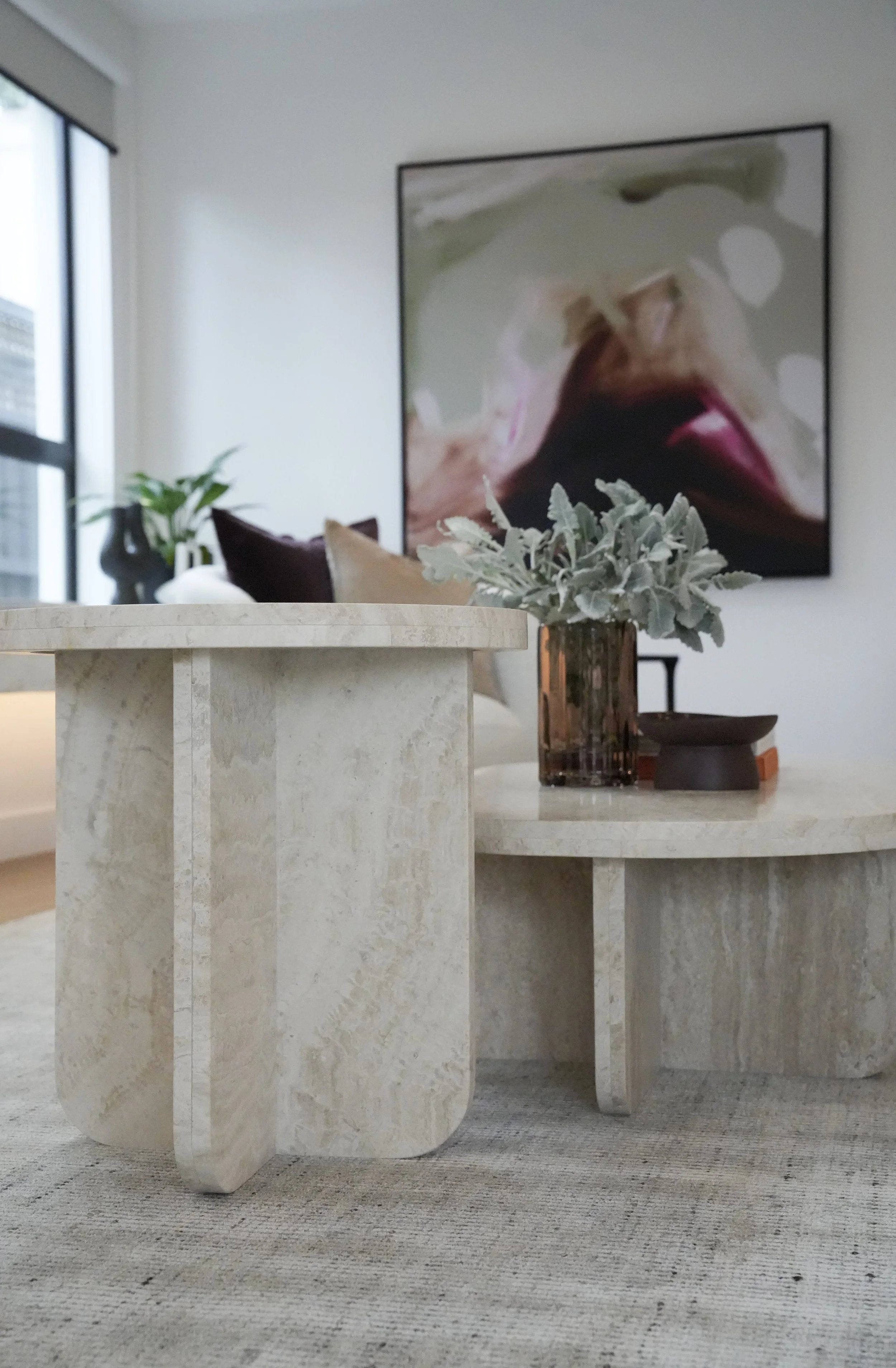 Modern living room with beige stone coffee tables, a brown vase with green plants, a large abstract painting on the wall, and a window with a cityscape view.