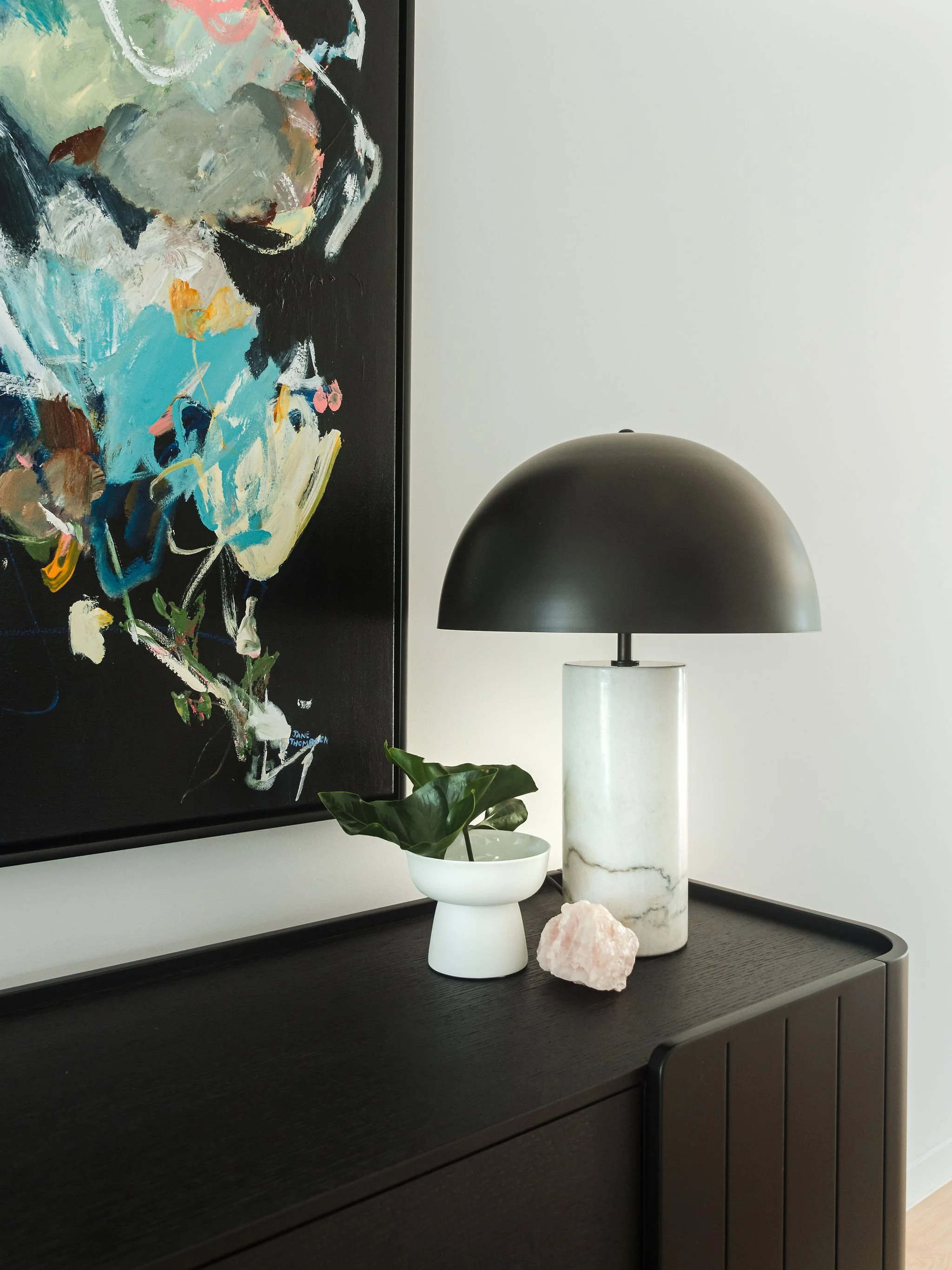 A modern interior decor scene featuring an abstract painting, a black dome-shaped table lamp with a marble base, a potted plant with dark green leaves in a white vase, and a pink salt rock on top of a black surface.