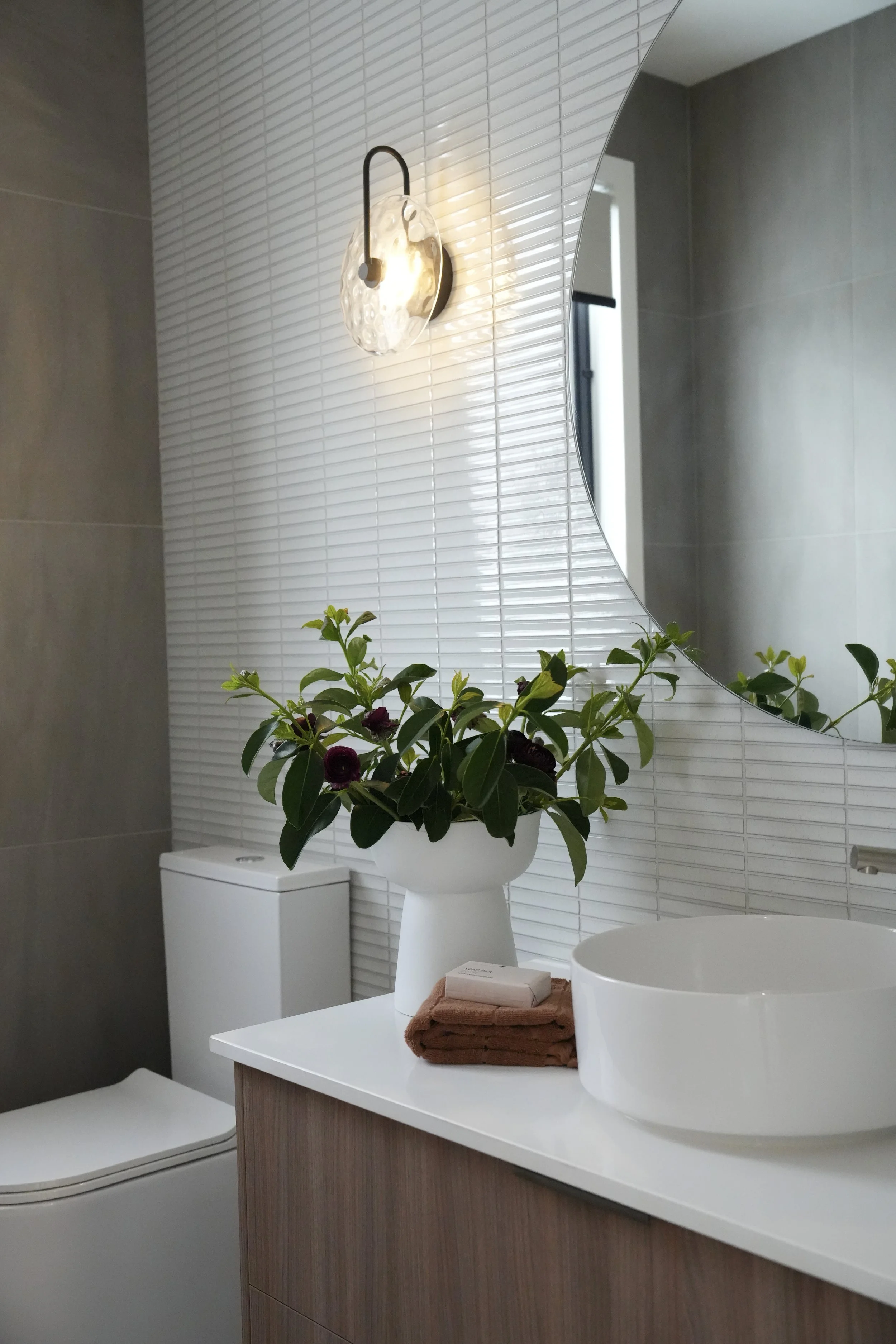 Modern bathroom with white countertop, round vessel sink, large oval mirror, a white vase with greenery and purple flowers, a folded brown towel, and a soap, with a wall-mounted light fixture and a toilet in the background.