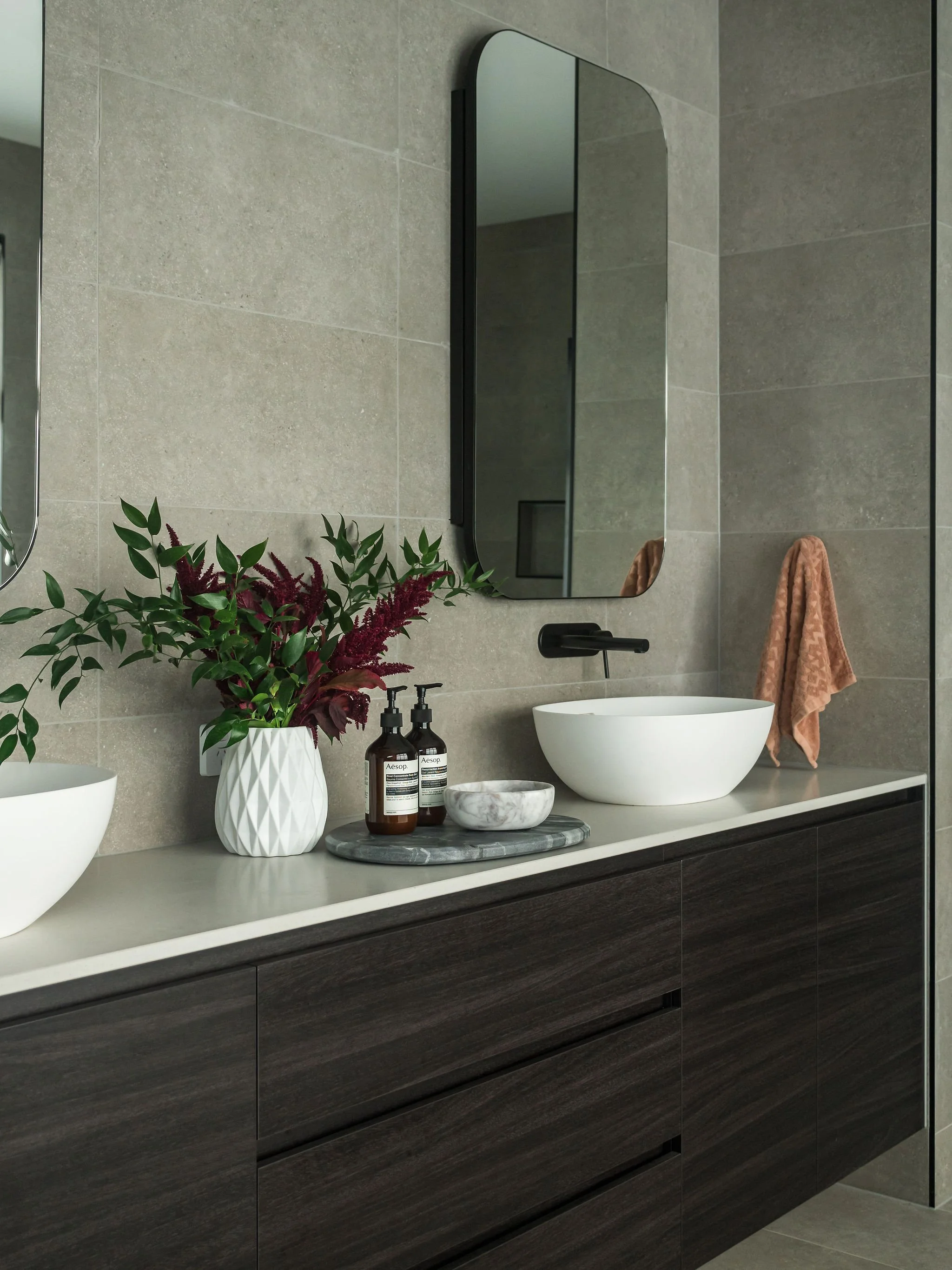 Modern bathroom vanity with a large mirror, a white vessel sink, dark wood cabinetry, a marble tray with soap dispensers, and a vase with greenery, beige tiled wall, and a peach-colored towel hanging on the wall.