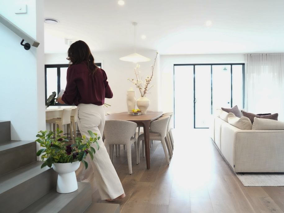 Woman standing on stairs in a modern, well-lit living and dining area with white walls, a large window, a beige sofa, a round dining table with chairs, decorative vases with branches, and a potted plant.