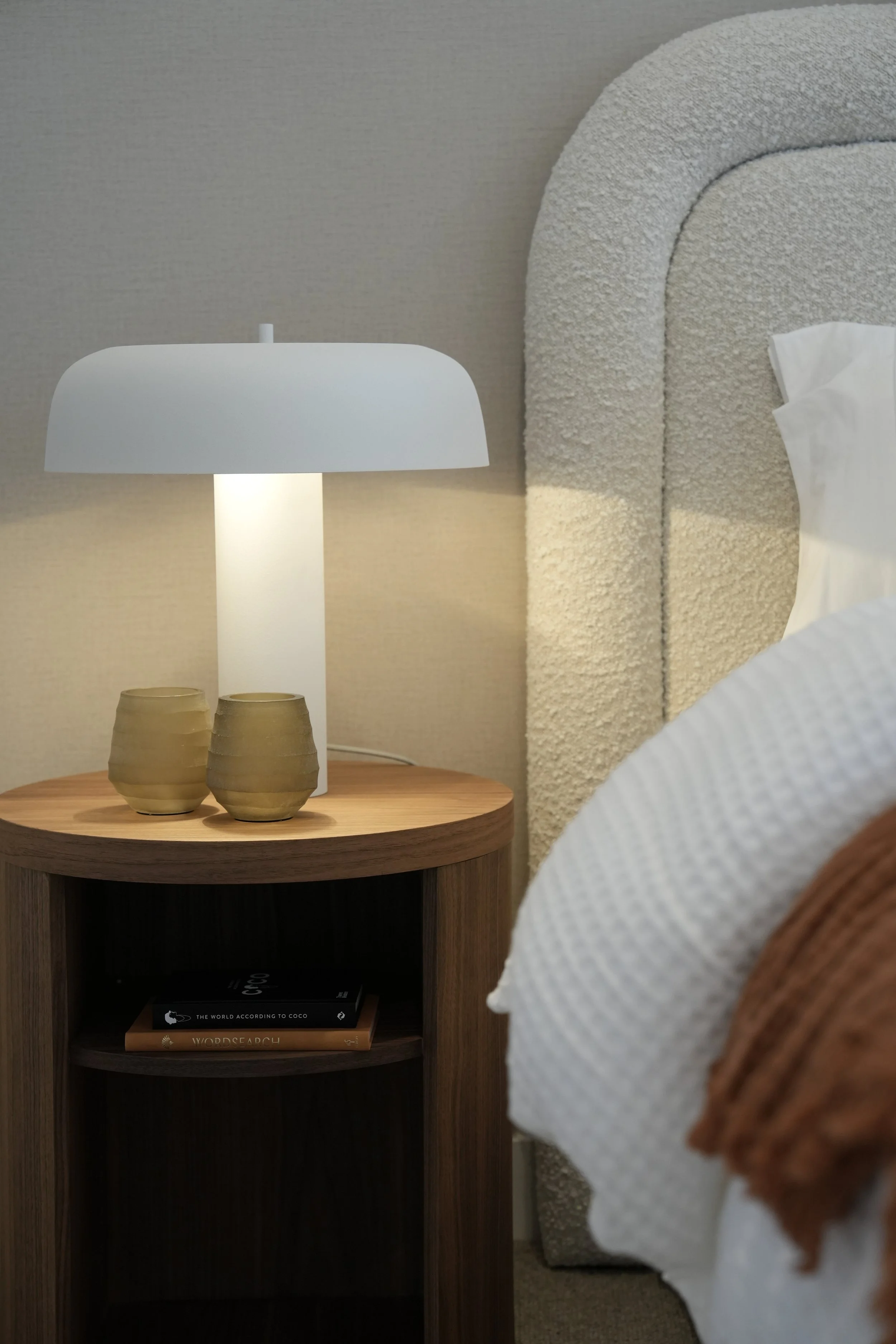 Close-up of a bedside table with a white lamp, two yellowish-green ceramic cups, and books underneath. Part of a bed with a curved, textured headboard and white bedding is visible.