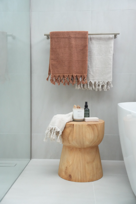 Bathroom with a wooden stool holding toiletries, a white towel, a wall-mounted towel rack with a peach-colored and a white towel, and a white bathtub.
