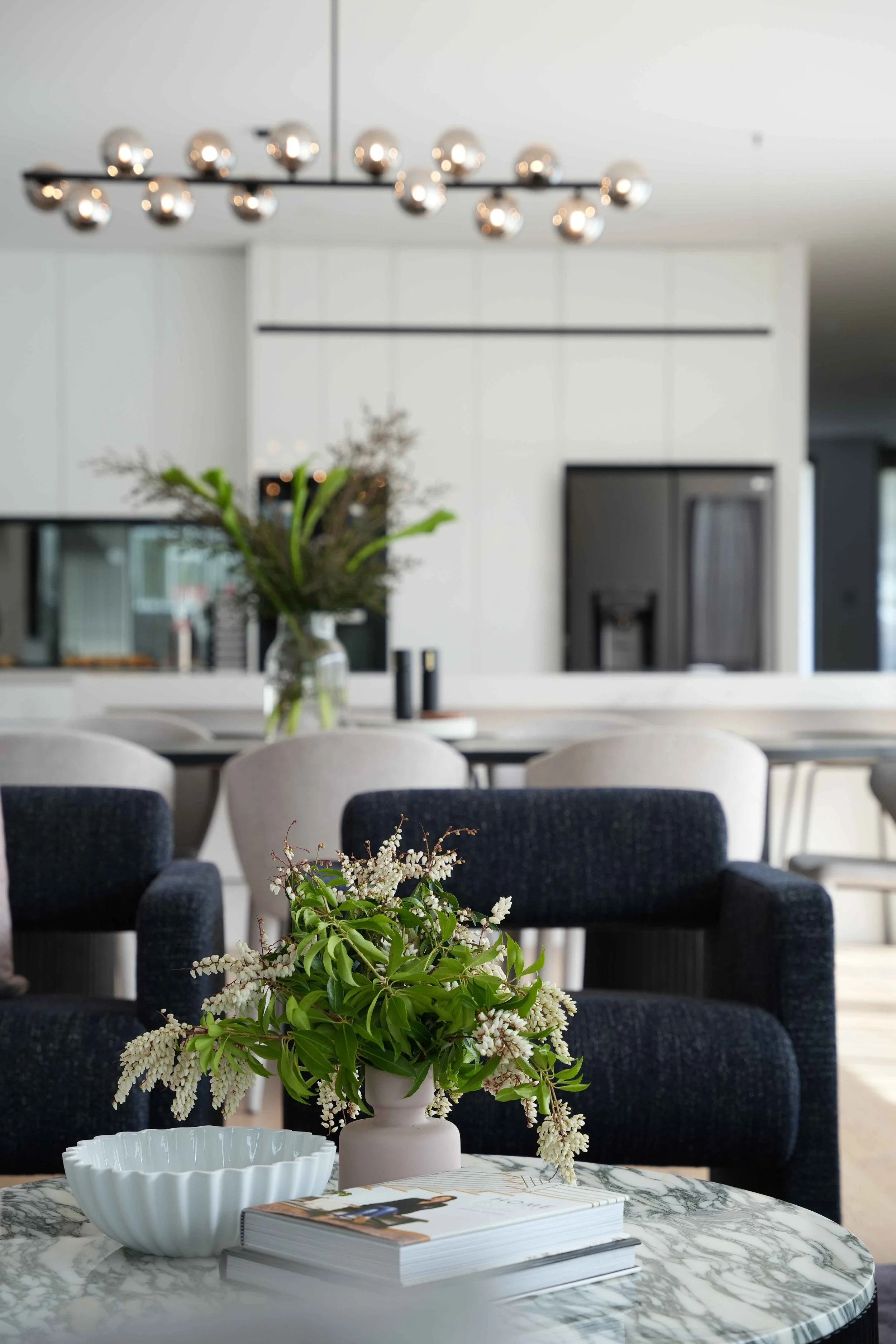 A living room with a marble coffee table, a white vase with white and green flowers, a bowl, and magazines, with a black sofa in the background and a hanging modern chandelier.