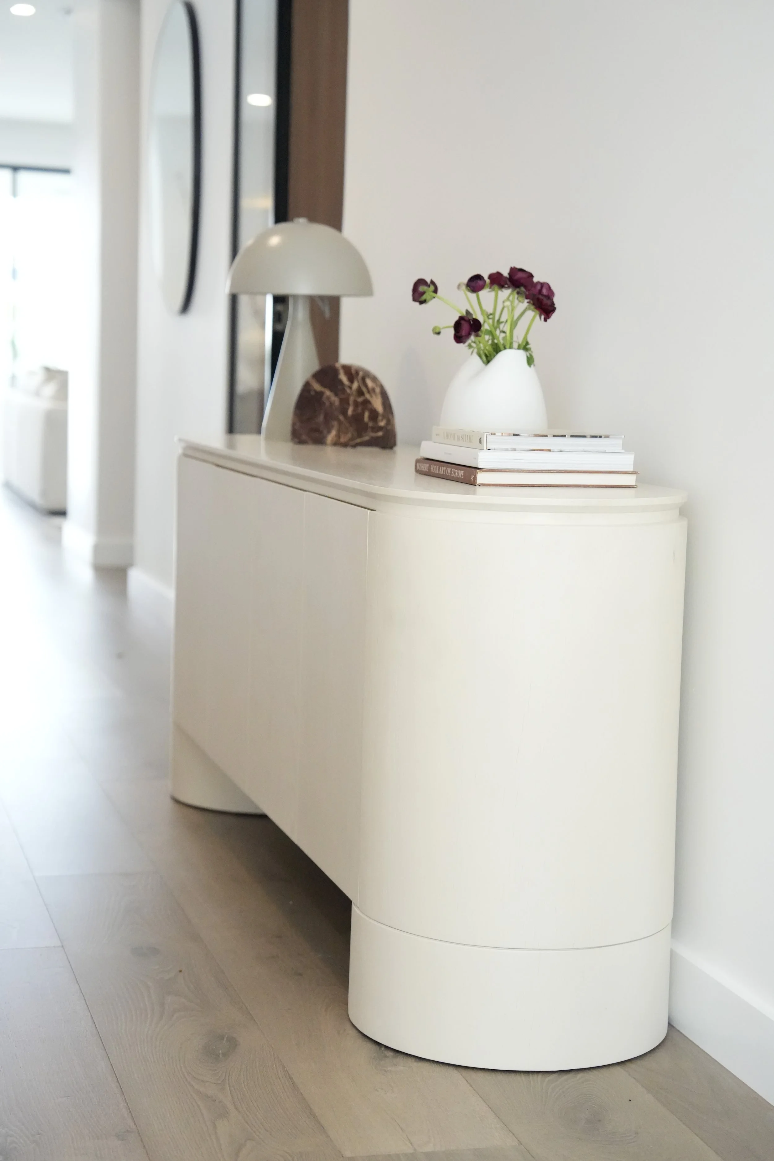 A white console table with a flower vase, a lamp, and some stacked magazines in a modern interior