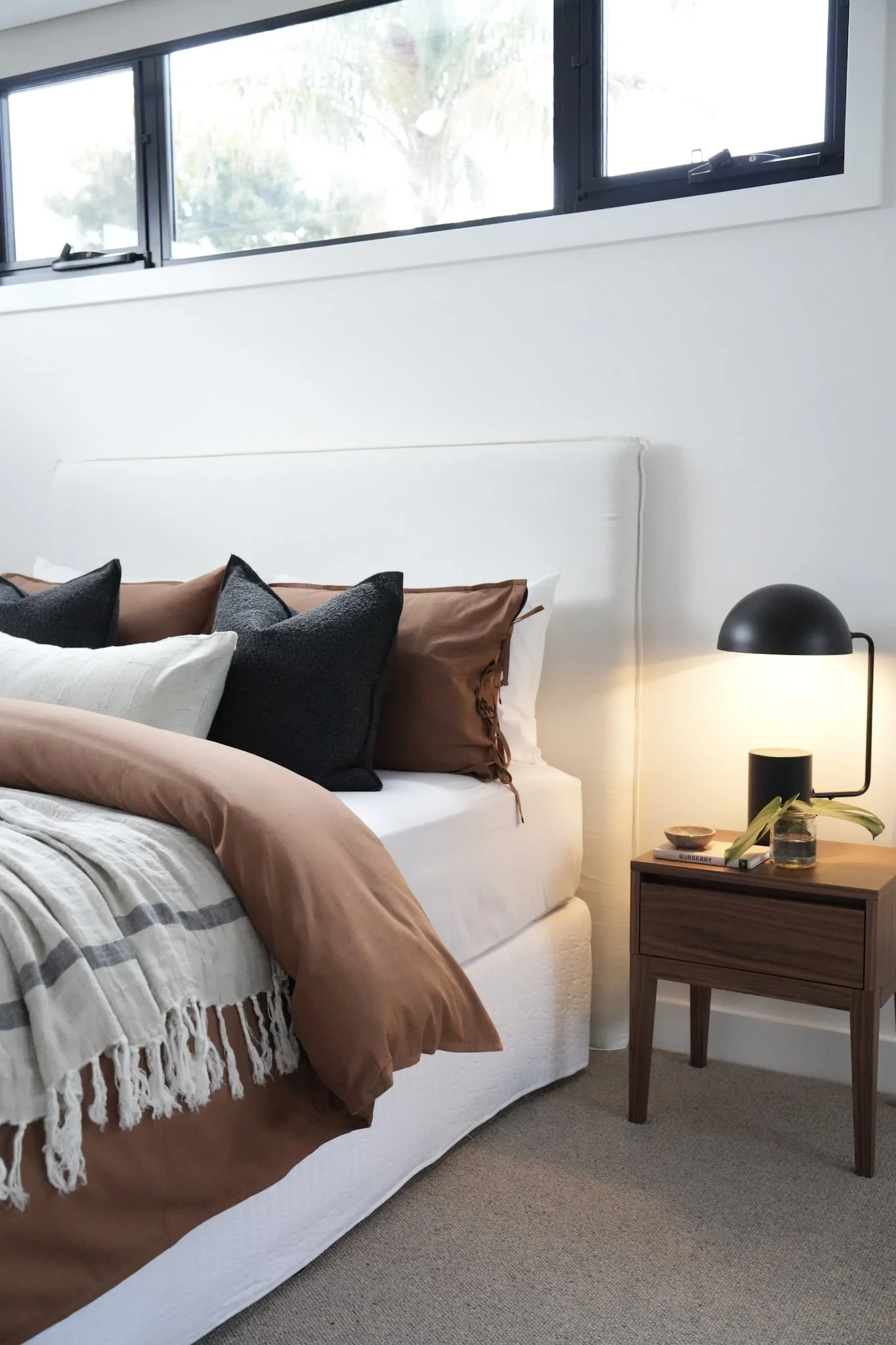 A neatly made bed with brown, white, and black pillows and a beige throw blanket in a bedroom with a nightstand and a black desk lamp, near a window with a view of trees.