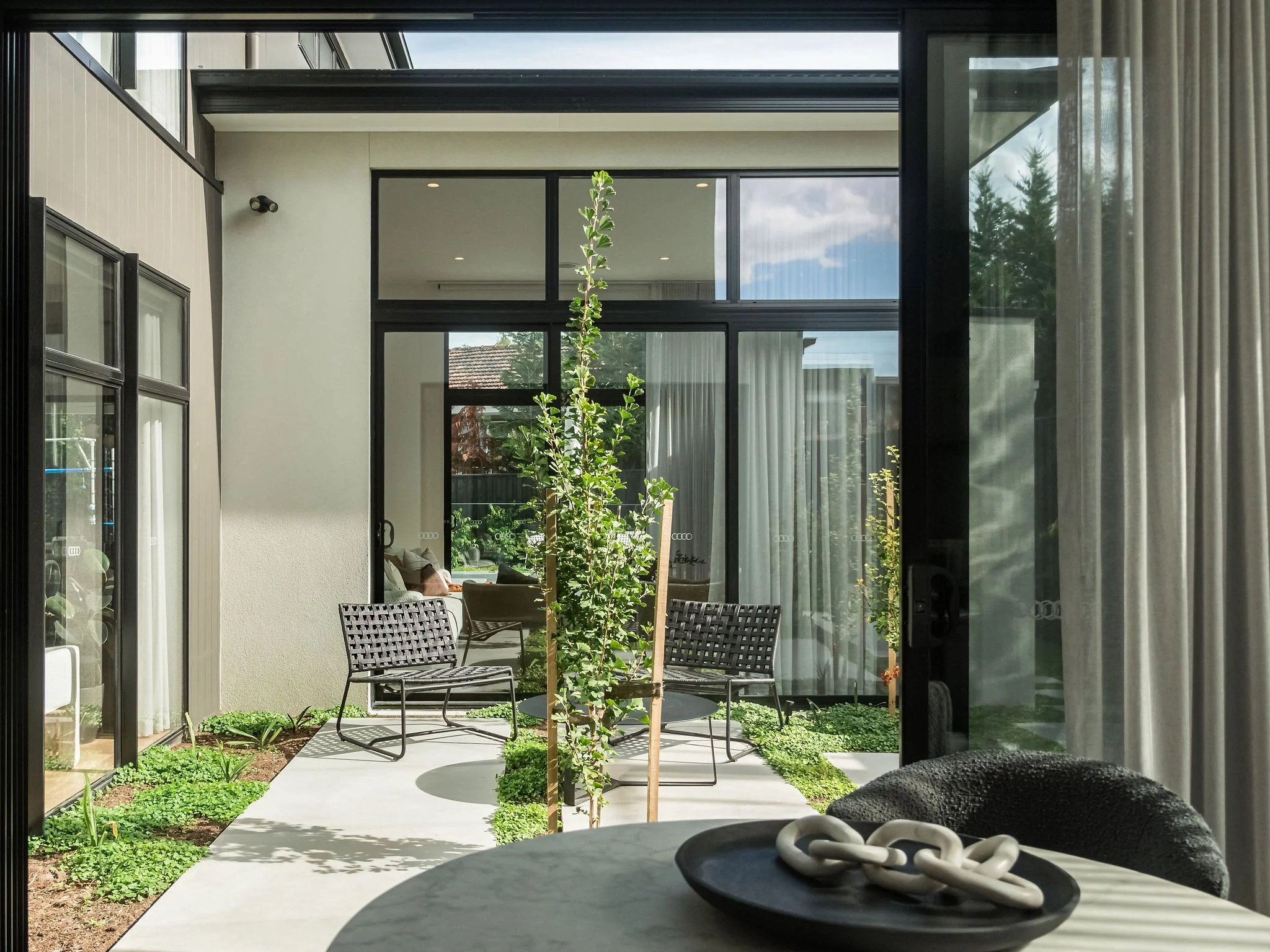 View of an indoor-outdoor living space with sliding glass doors, patio chairs, small trees, and a garden, seen from inside a home.