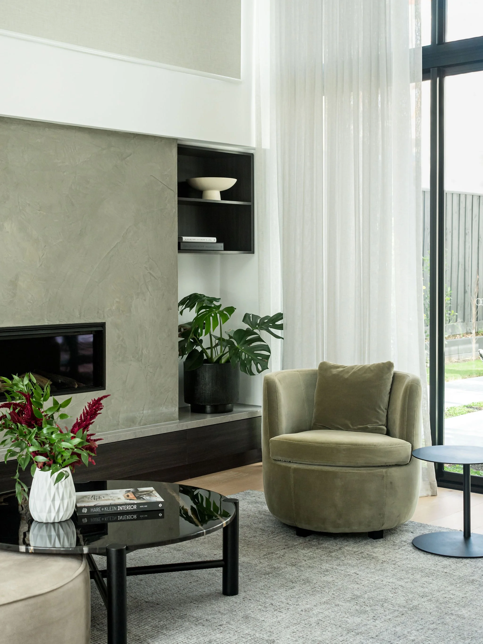 Modern living room with green armchair, black coffee table with books and flower vase, potted plant, fireplace, wall shelf, and large window with sheer curtains.