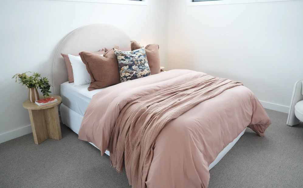 A neatly made bed with a pink duvet, multiple pillows including a floral pillow, and a small wooden side table holding a plant and a magazine in a bright, modern bedroom.