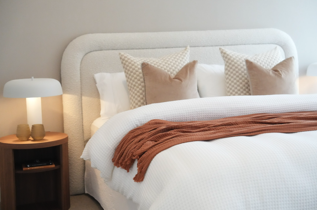 Well-made bed with white quilt, brown throw blanket, four decorative pillows, and a wooden nightstand with a lamp and two small vases.