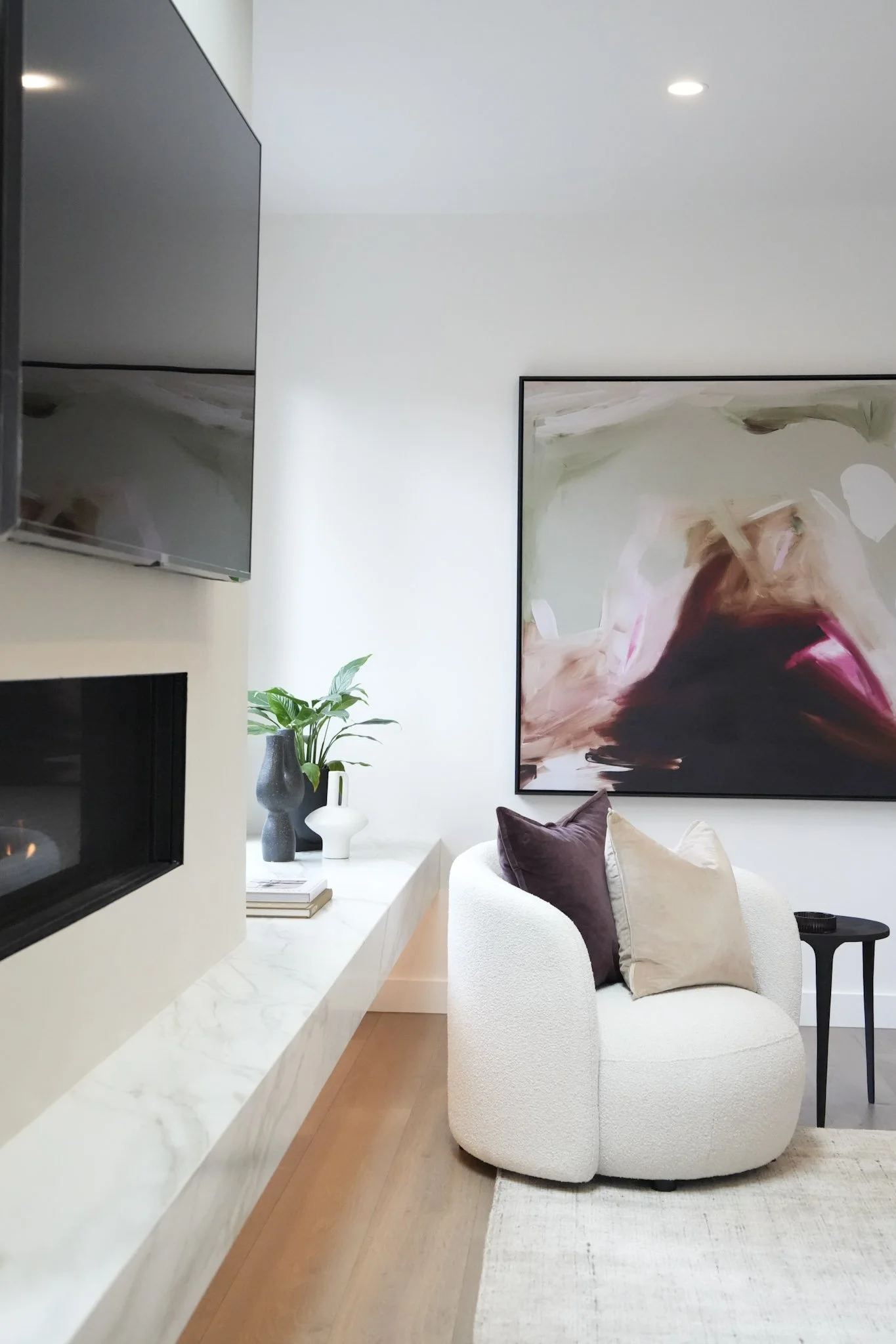 Modern living room corner with white textured armchair, purple and beige pillows, abstract colorful painting, potted plant, fireplace, wall-mounted TV, side table, and wooden floor.