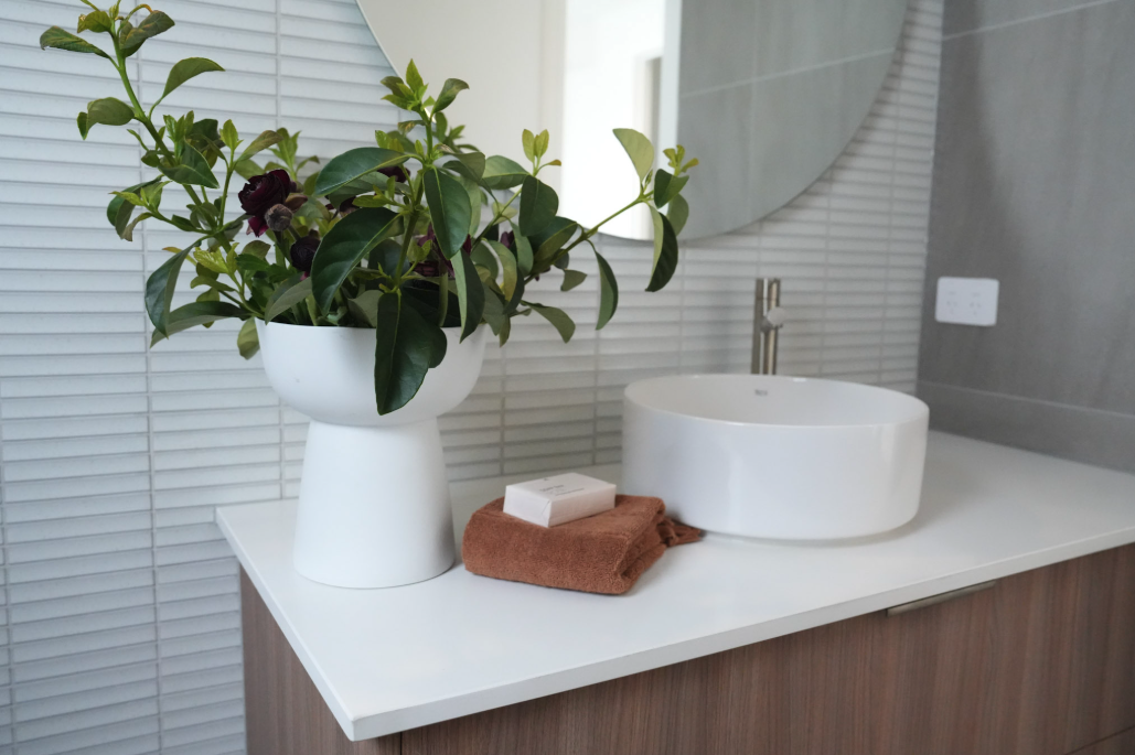Bathroom countertop with a white vase holding green foliage, a bar of soap, and a brown towel beside a white vessel sink, with a round mirror in the background.