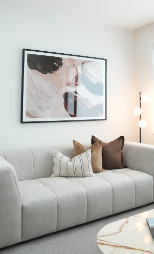 Living room with a light gray sofa decorated with striped and solid-colored pillows, a framed abstract art piece on the wall, and a modern floor lamp with three round lights.