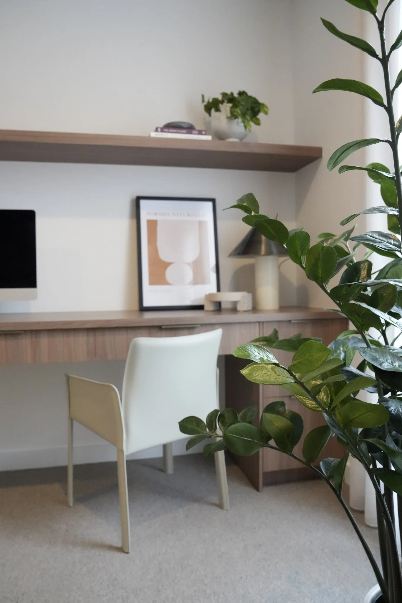 A home office with a desk, white chair, and houseplant. The desk has a framed abstract art print, a lamp, and a small decorative item.