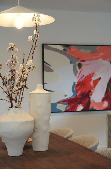 Interior dining room with a wooden table holding two white vases with cherry blossom branches, a modern pendant light overhead, and abstract artwork on the wall.