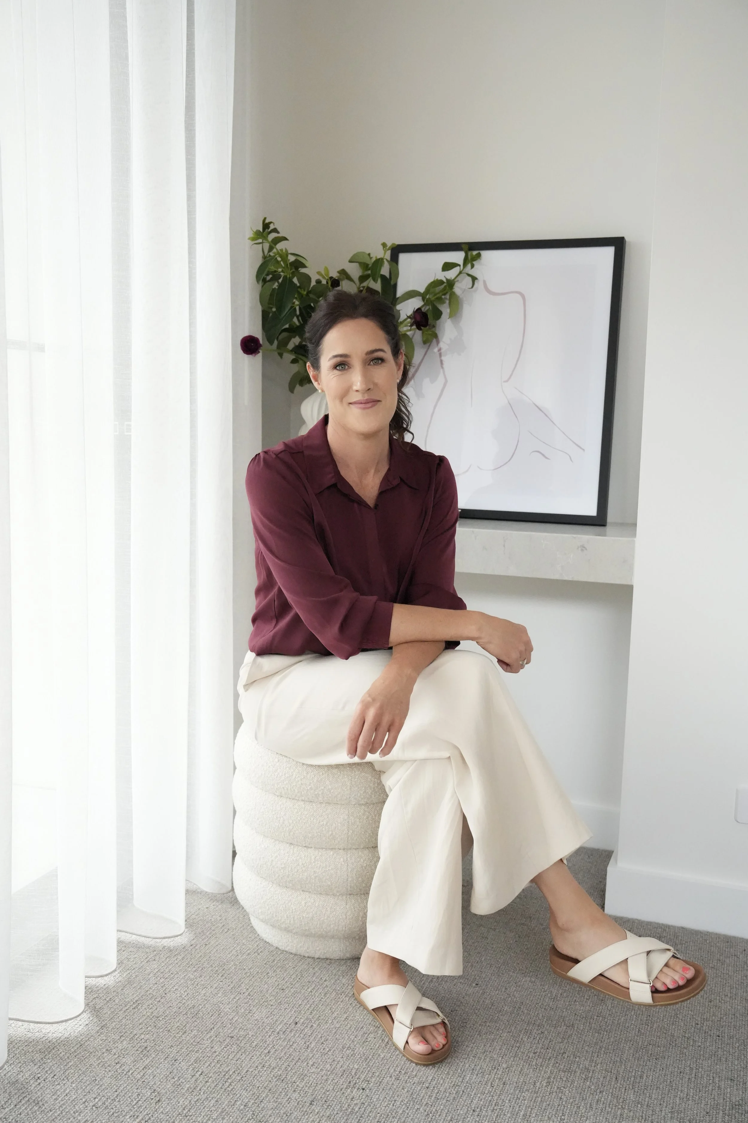 A woman with dark hair, wearing a burgundy blouse and white wide-leg pants, sits on a textured white stool in a bright, modern room. She has her arms crossed and is smiling slightly at the camera. Behind her is a framed abstract line drawing leaning 