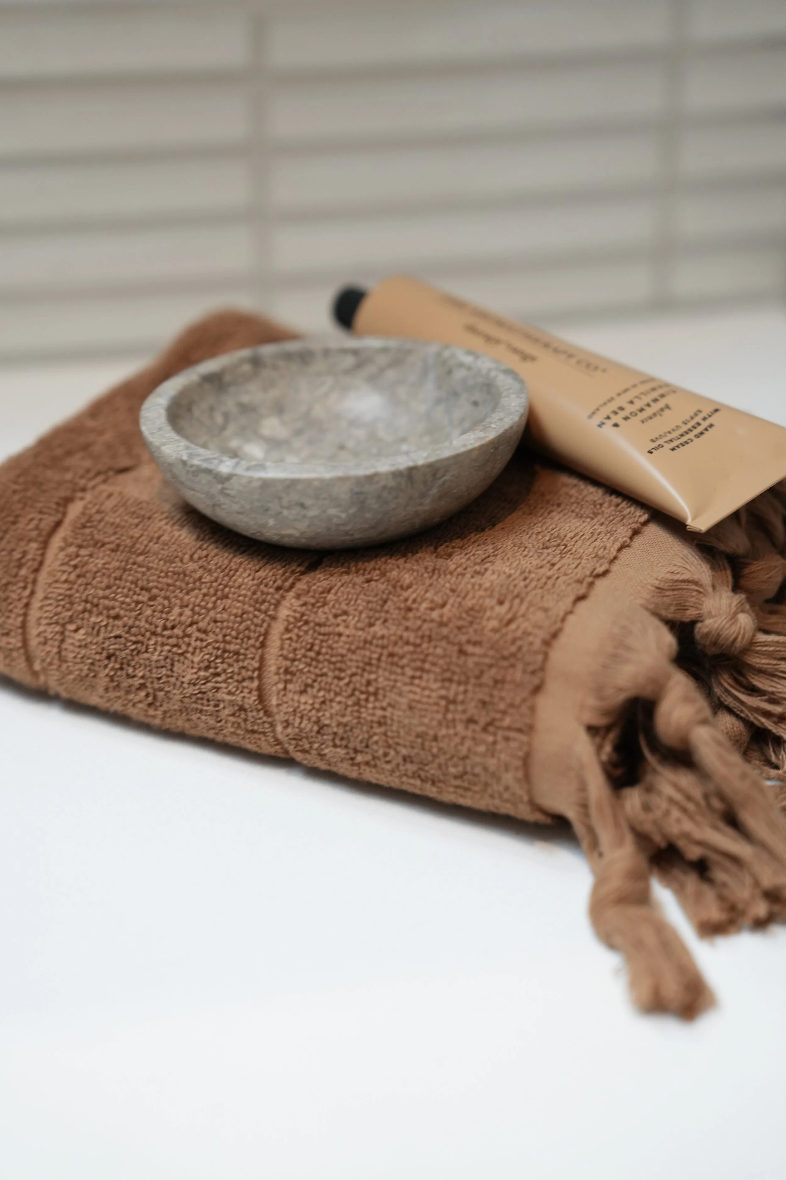 A rolled brown towel with a small gray stone bowl and a beige tube of skincare product on top, placed against a white background.