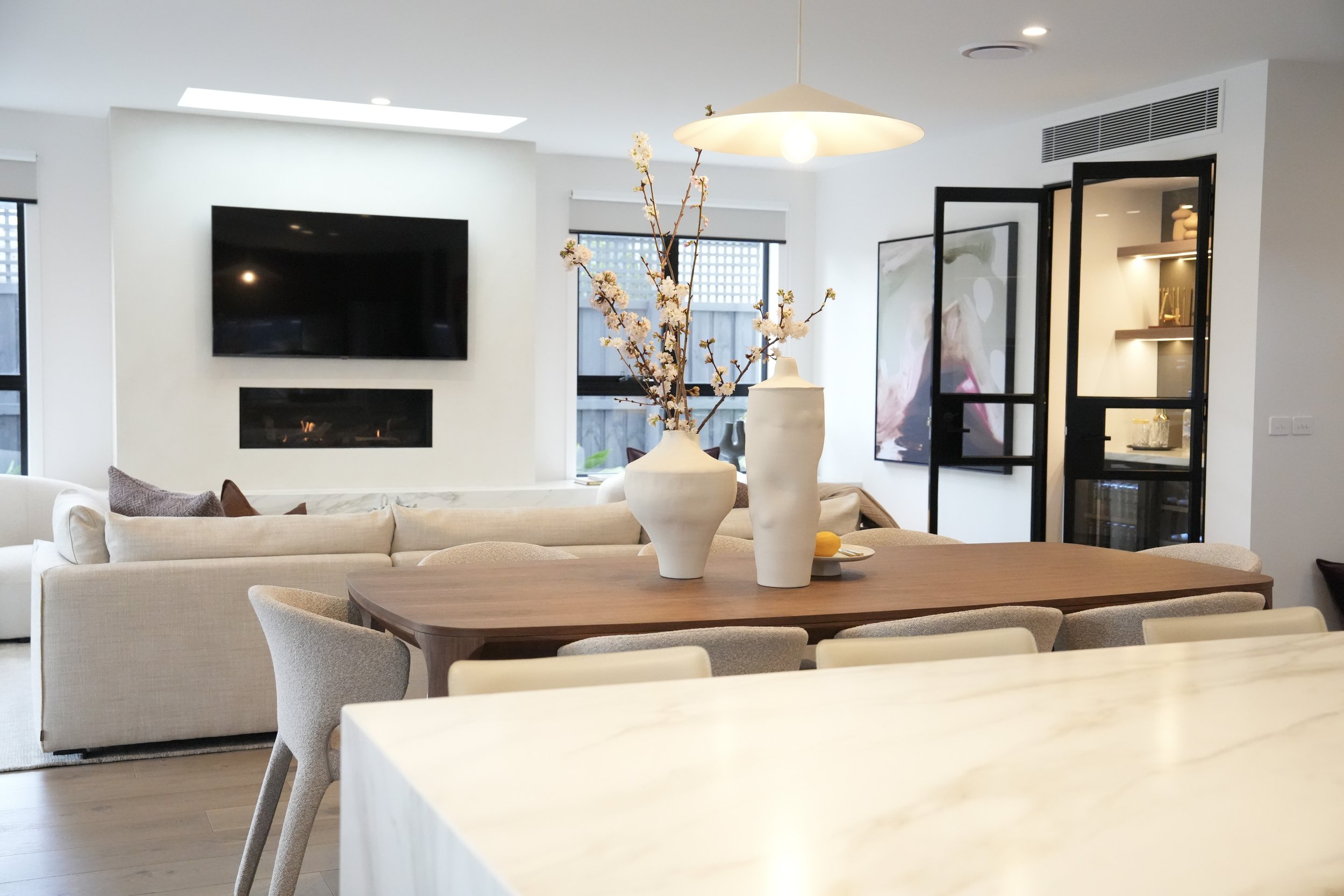Modern living room with a wooden dining table, two white vases with pink flowers, beige upholstered chairs, a cream-colored sofa, a wall-mounted flat-screen TV, and a fireplace.