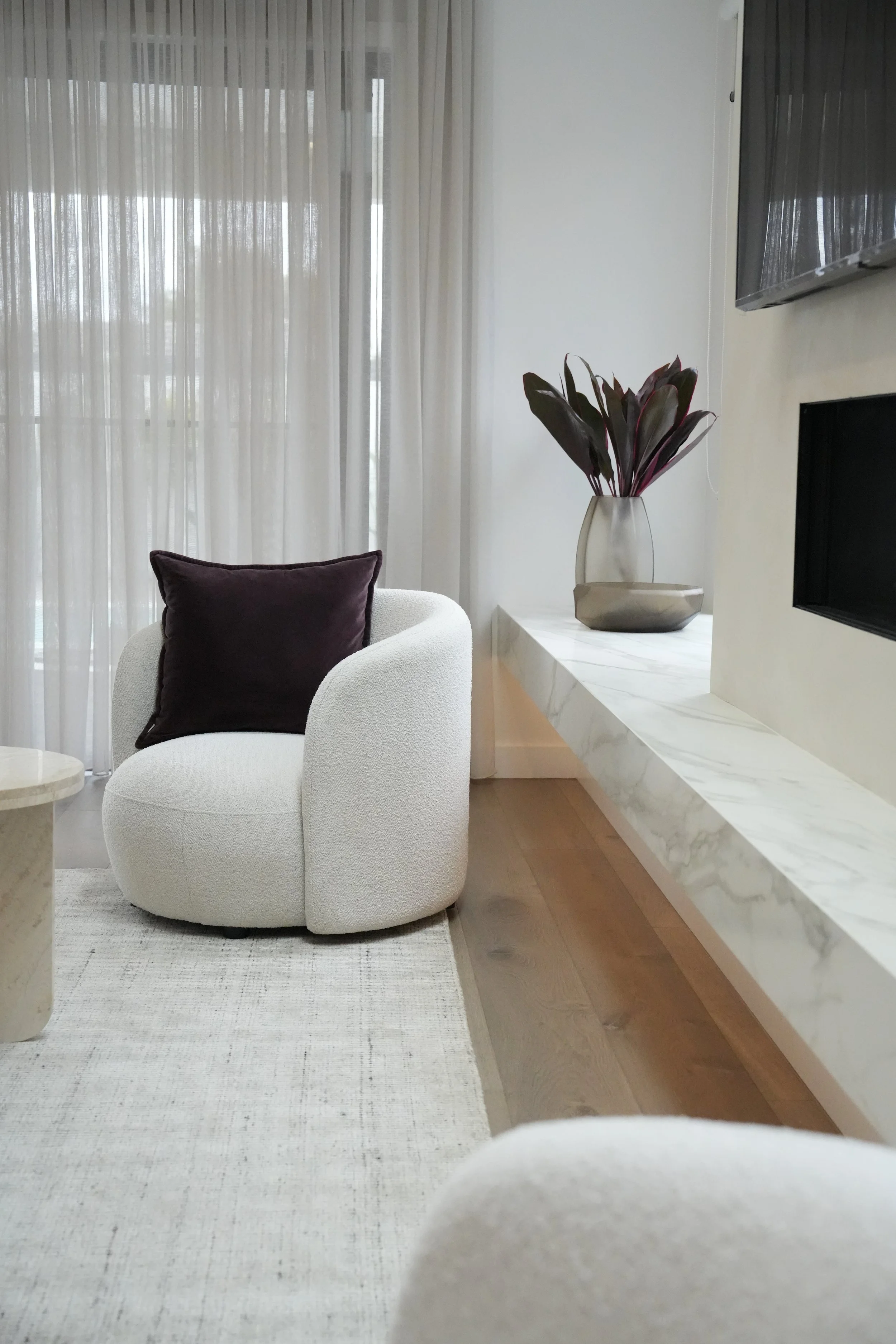 A modern living room corner with a white upholstered armchair with a dark purple pillow, a marble side table, a marble shelf with a vase of dark purple leaves, beige walls, and sheer curtains covering a window.