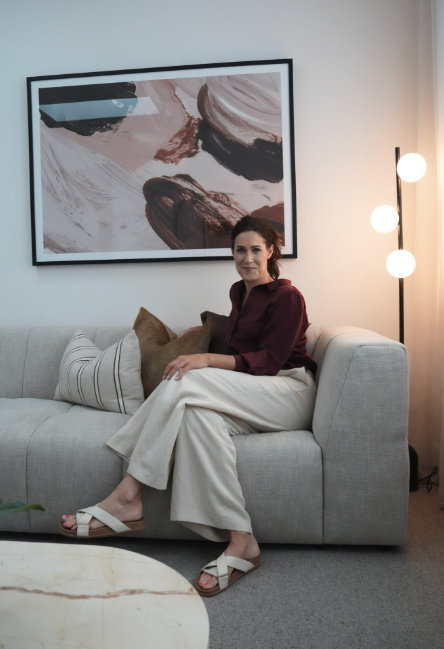 A woman sitting on a light gray sofa with beige pillows in a modern living room, with a large abstract painting on the wall and a floor lamp with three globe lights beside her.