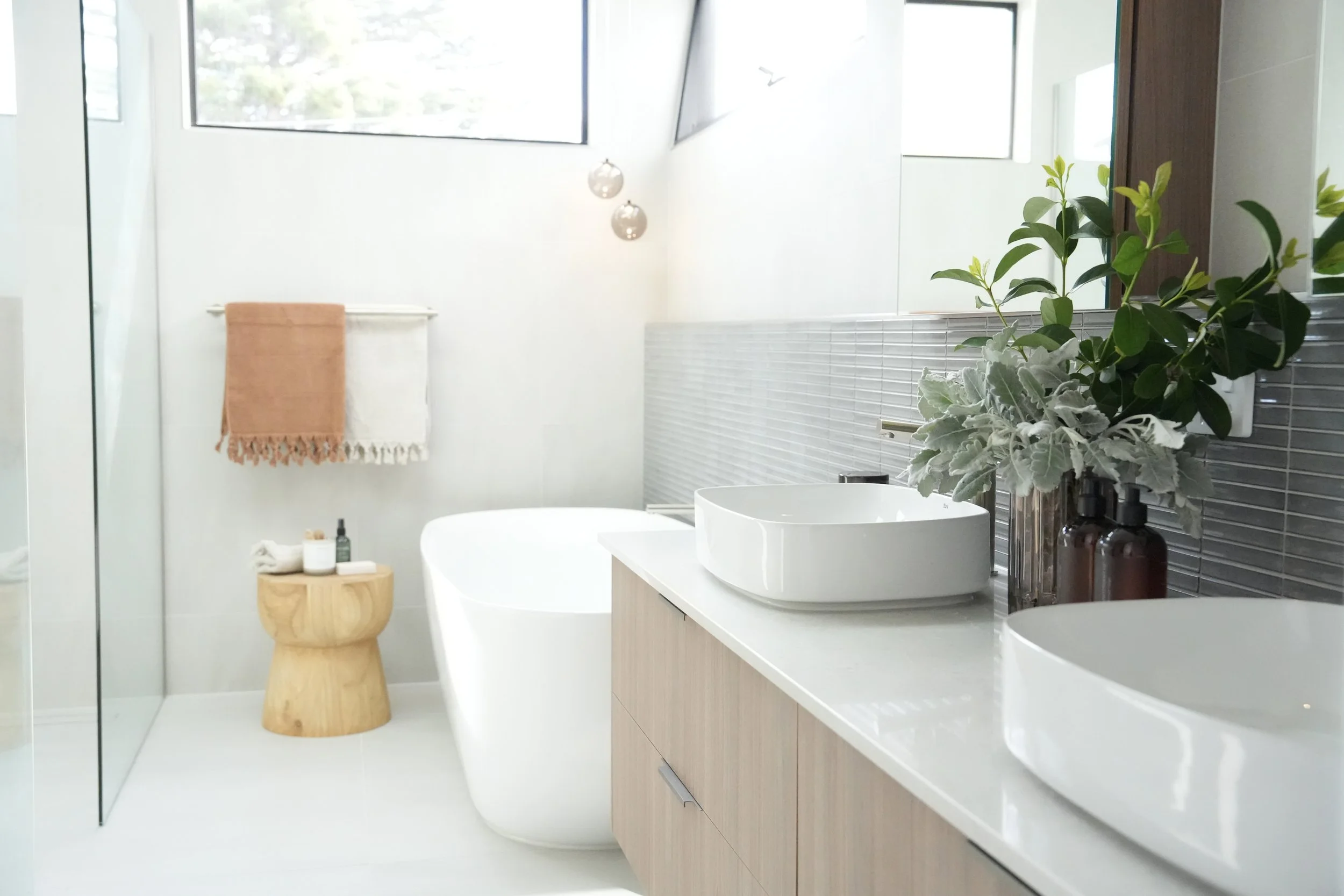 Modern bathroom with white walls, large window, double vessel sinks with brown soap dispensers, decorative plant, bathtub, wooden stool with toiletries, and beige towels on a towel rack.