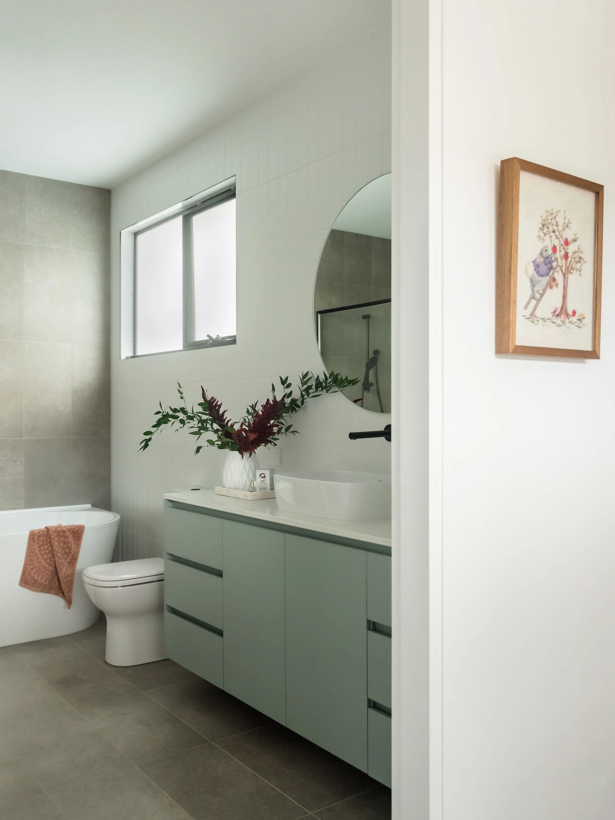 Modern bathroom with a bathtub, floating teal vanity with drawers, round mirror, frosted window, and floral arrangements. There is a framed art piece on the wall.