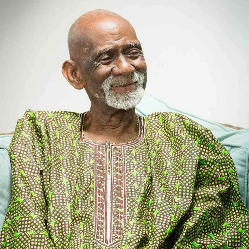 An elderly man with a bald head and white beard, sitting on a light blue couch, smiling and wearing a green patterned shirt.