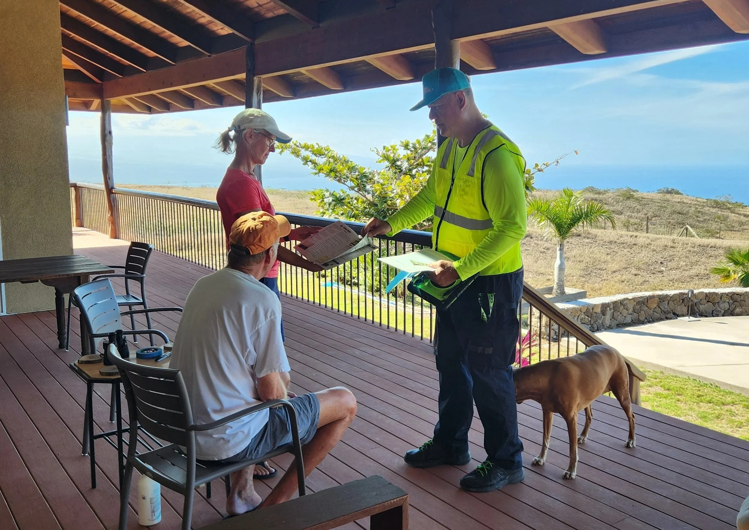Three people and a dog on a covered porch overlooking a landscape with grass, trees, and the ocean in the distance. One person is sitting, another is standing with papers, and the third is also sitting and facing away. The dog stands nearby.