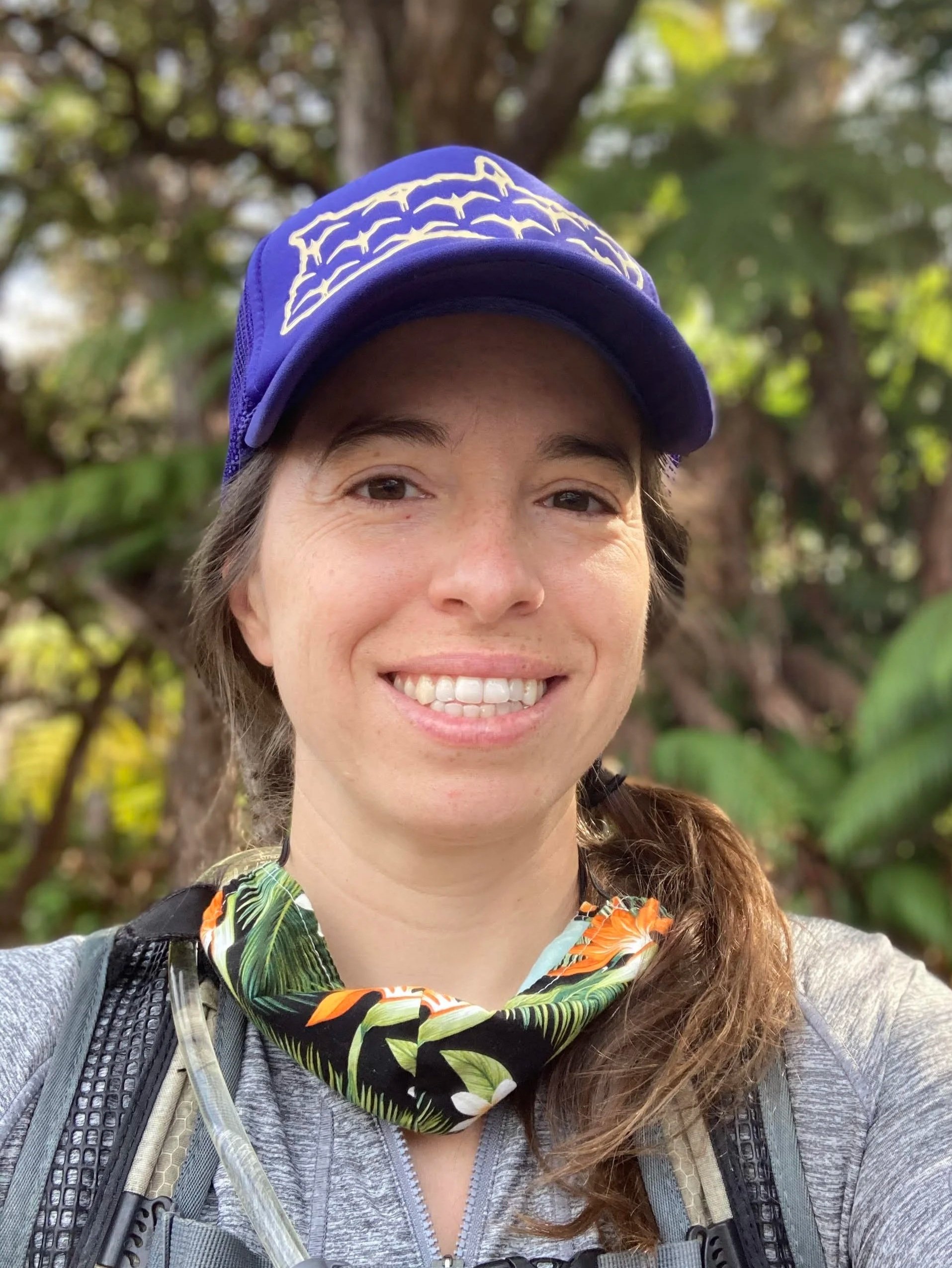 A woman smiling outdoors with trees and green foliage in the background, wearing a purple cap, a gray athletic jacket, a patterned neck gaiter, and a backpack.