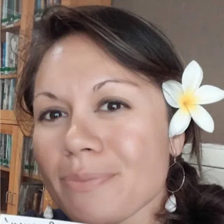A woman with dark hair and a white flower in her ear, smiling softly in an indoor setting with bookshelves in the background.