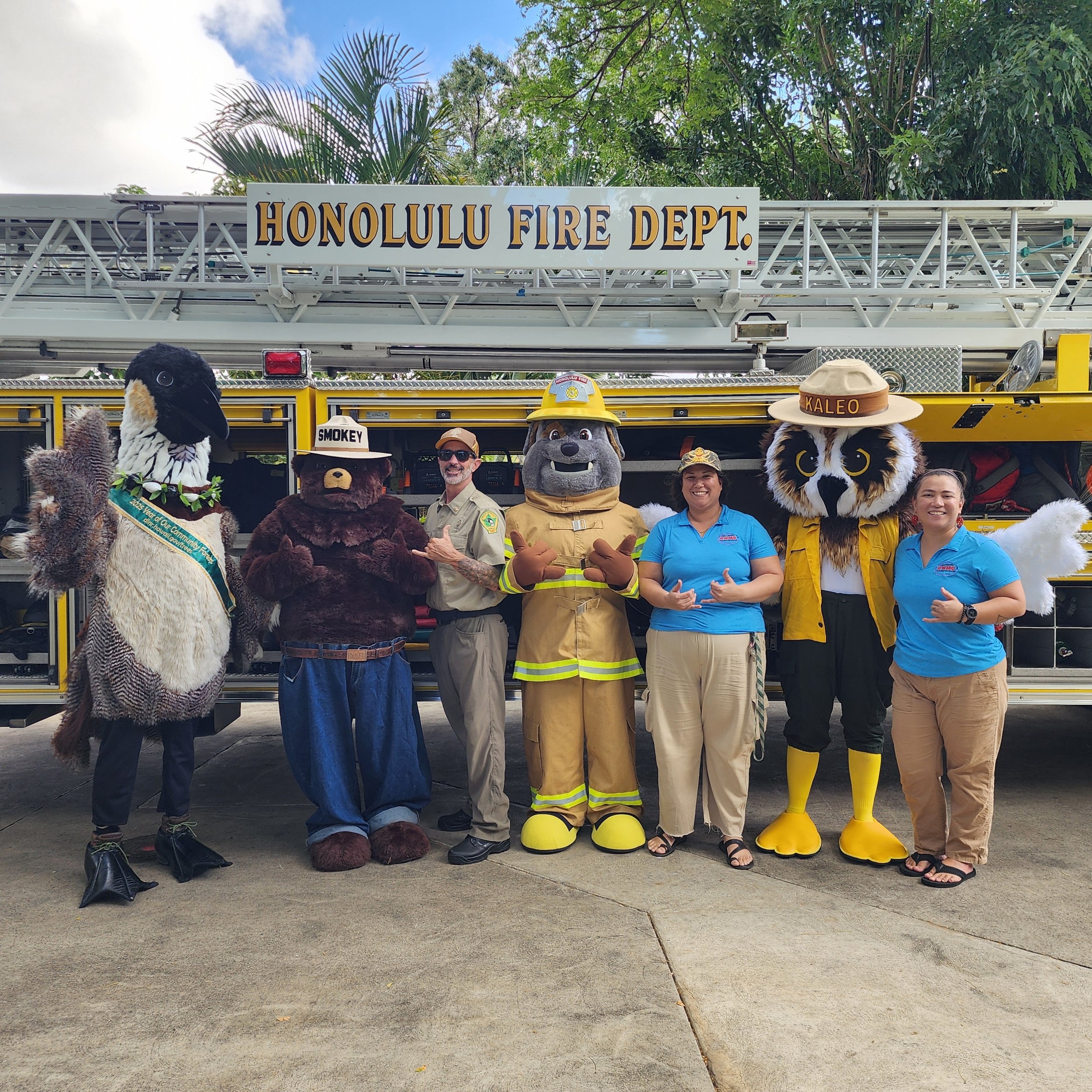 Group of people in costume and uniform standing in front of a fire truck labeled 'Honolulu Fire Dept.' with a large ladder on top. They are posing for a photo, some smiling and making hand gestures, with trees and a blue sky in the background.