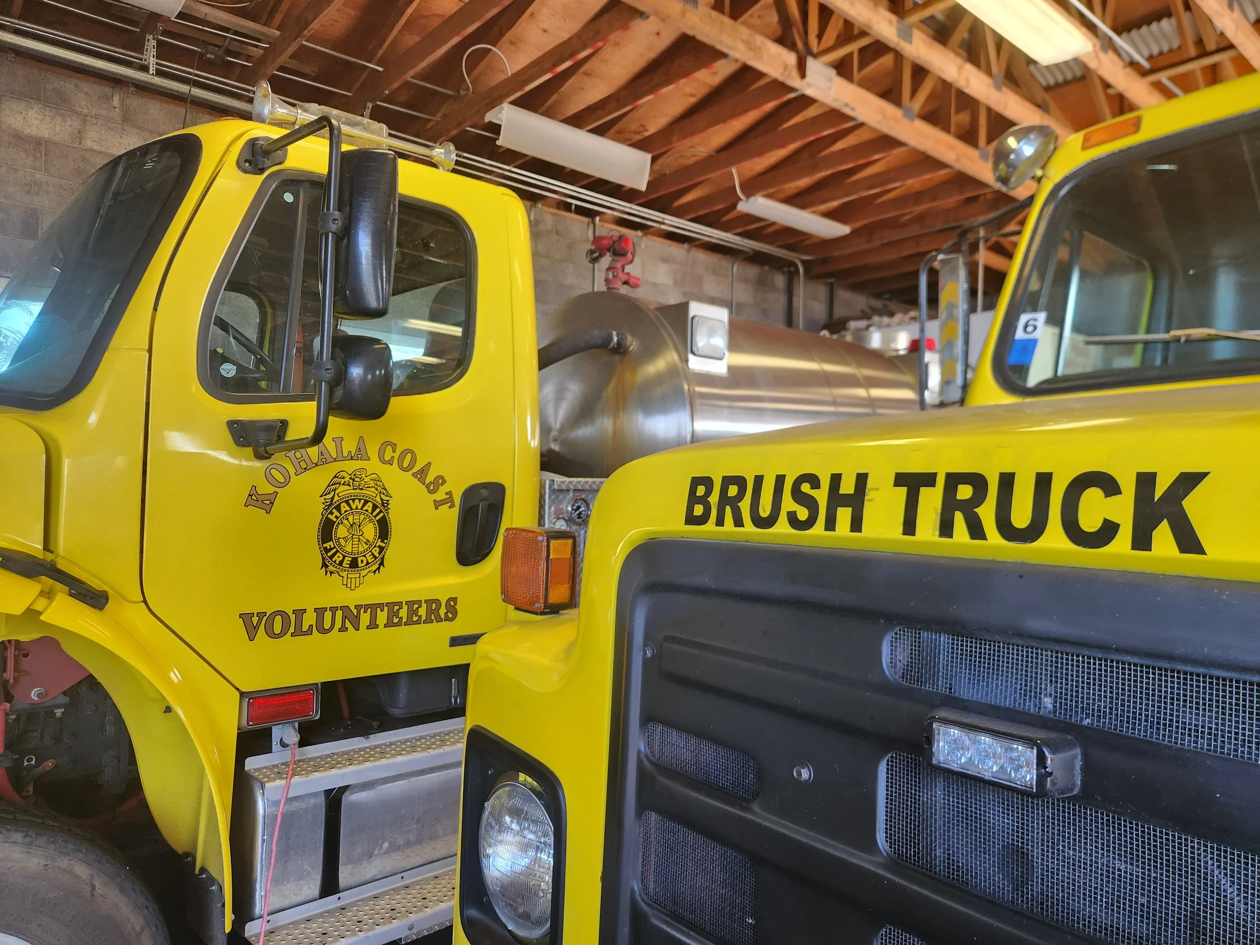 Yellow fire trucks marked 'Kohala Coast Volunteers' and 'Brush Truck' parked inside a garage with wooden beams and fluorescent lights.