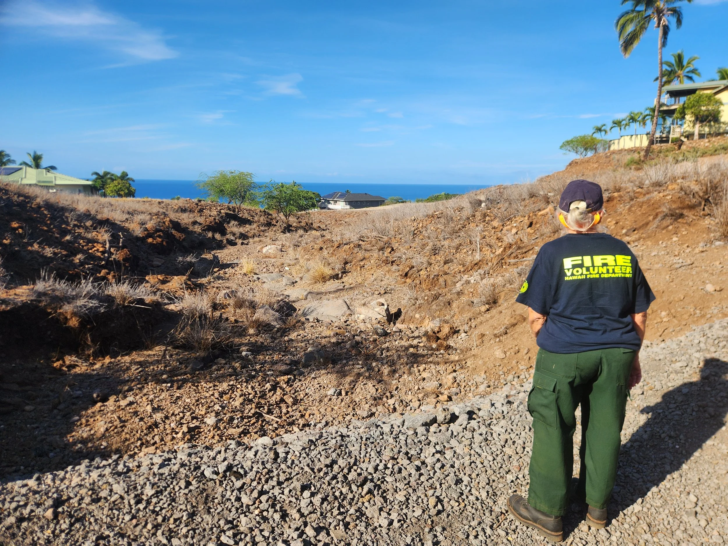 A person in a black Hawaii Fire Department fire volunteer shirt and green pants standing on a rocky, brown hillside with a view of the ocean and palm trees in the distance.